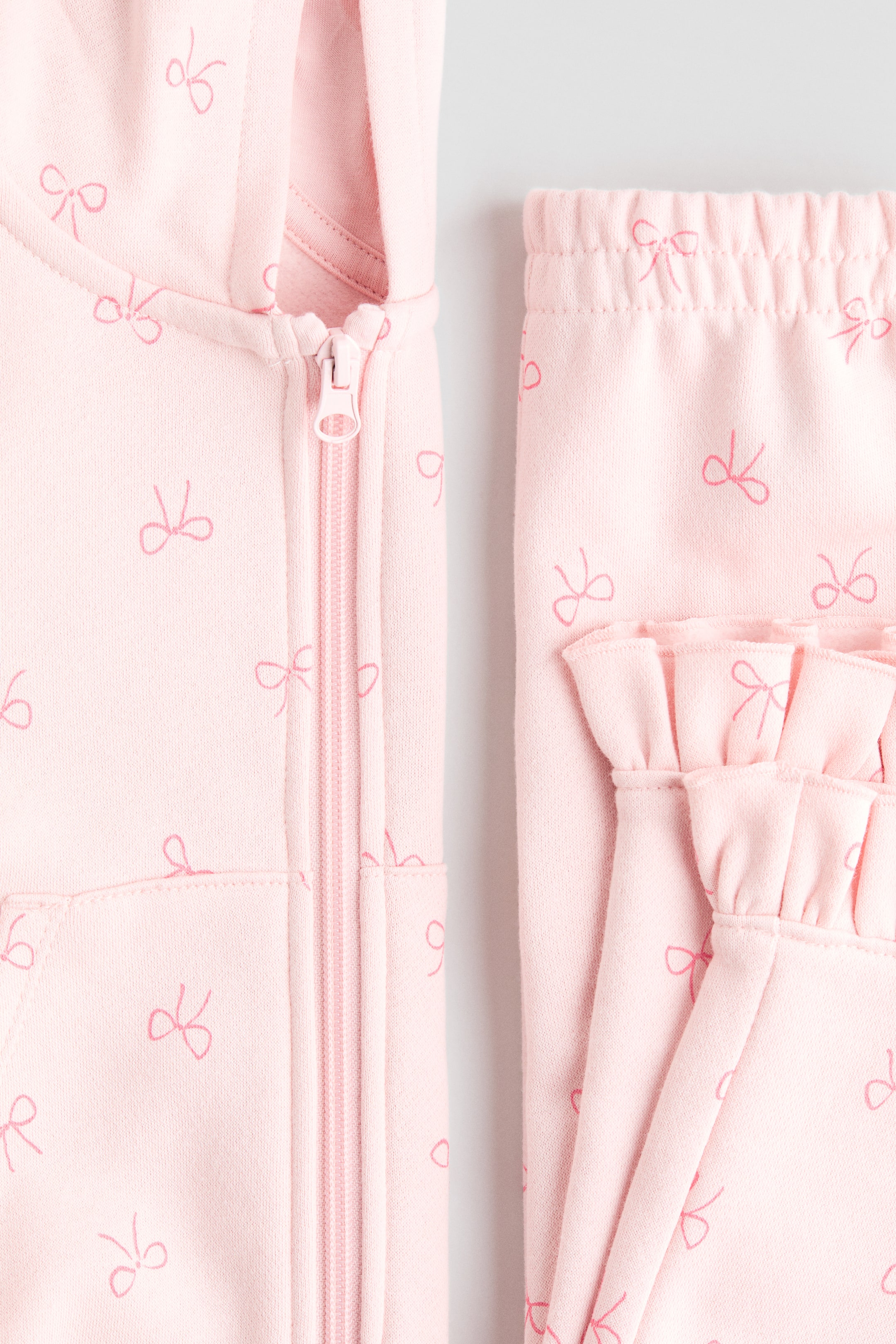 View larger image: A pink children's clothing set is being presented, featuring a hooded top with a full-length front zipper and a pocket, and matching bottoms. Both pieces are displaying a repeating pattern of small pink bow outlines, and the bottoms are showcasing an elasticated waistband and a ruffled cuff.
