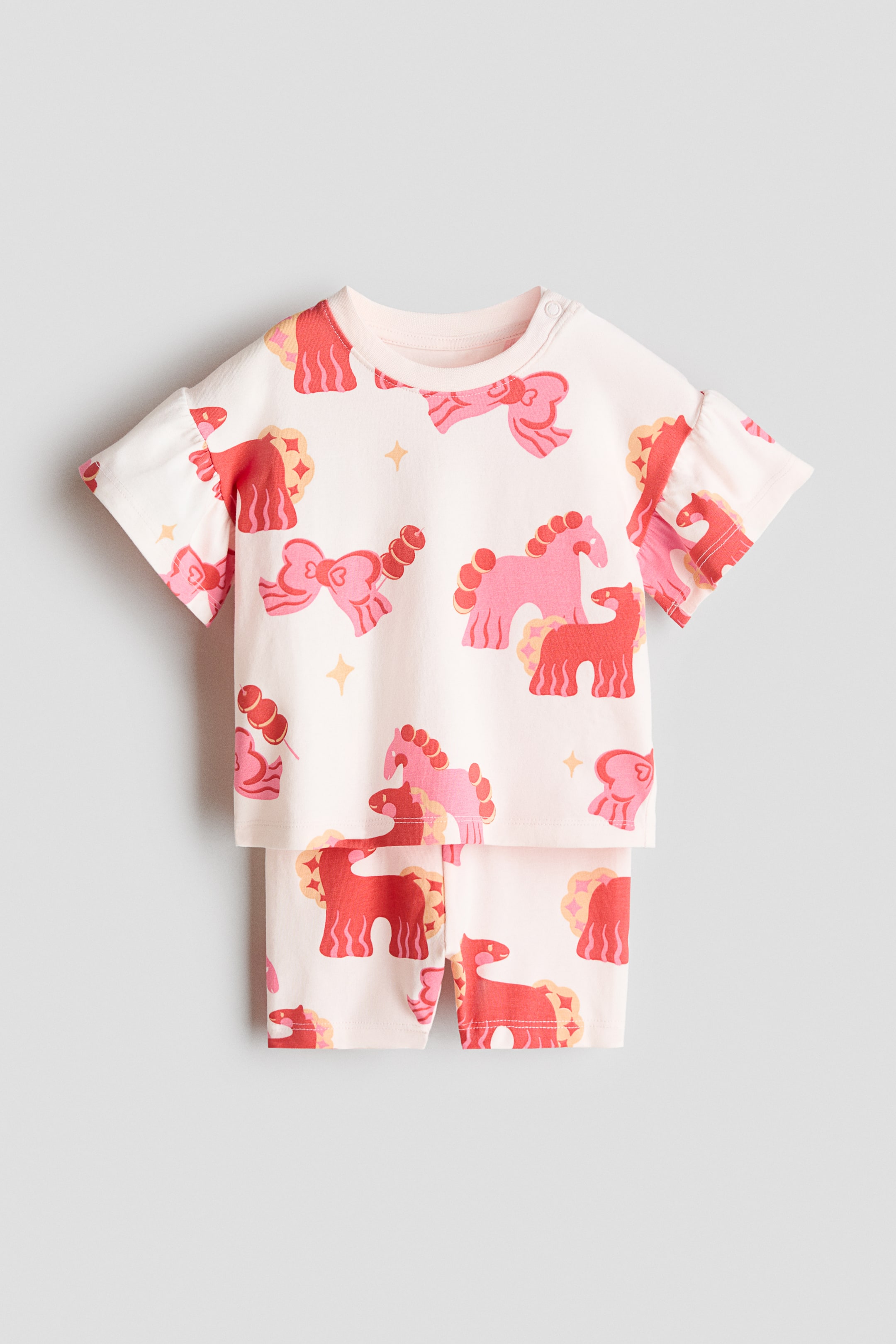 View larger image: A two-piece baby outfit is displaying a cream base with a repeating pattern of pink and red horses, pink bows, and small yellow stars, FRONT facing. The short-sleeved top is featuring ruffled sleeves and a round neck with two snap buttons on the shoulder, and matching shorts are forming the bottom piece.