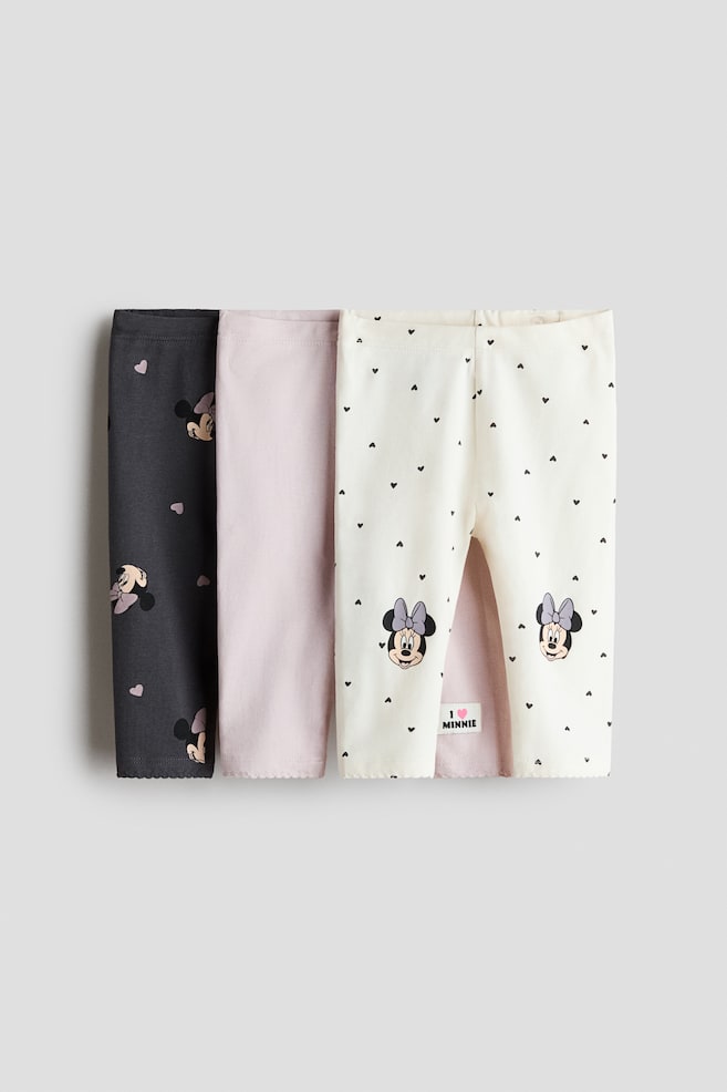 Drei Paar Baby-Leggings aus Baumwolljersey liegen flach in einer Ã¼bereinander angeordneten Darstellung und sind von vorne zu sehen. Das vordere Paar ist cremefarben und mit einem Allover-Print aus schwarzen Herzen sowie Minnie-Mouse-Grafiken an den unteren Beinen versehen, wÃ¤hrend die dahinter gefalteten Paare unifarben in Hellrosa und Dunkelgrau gehalten sind und mit einem pinken Herz- und Minnie-Mouse-Muster versehen sind. - 1