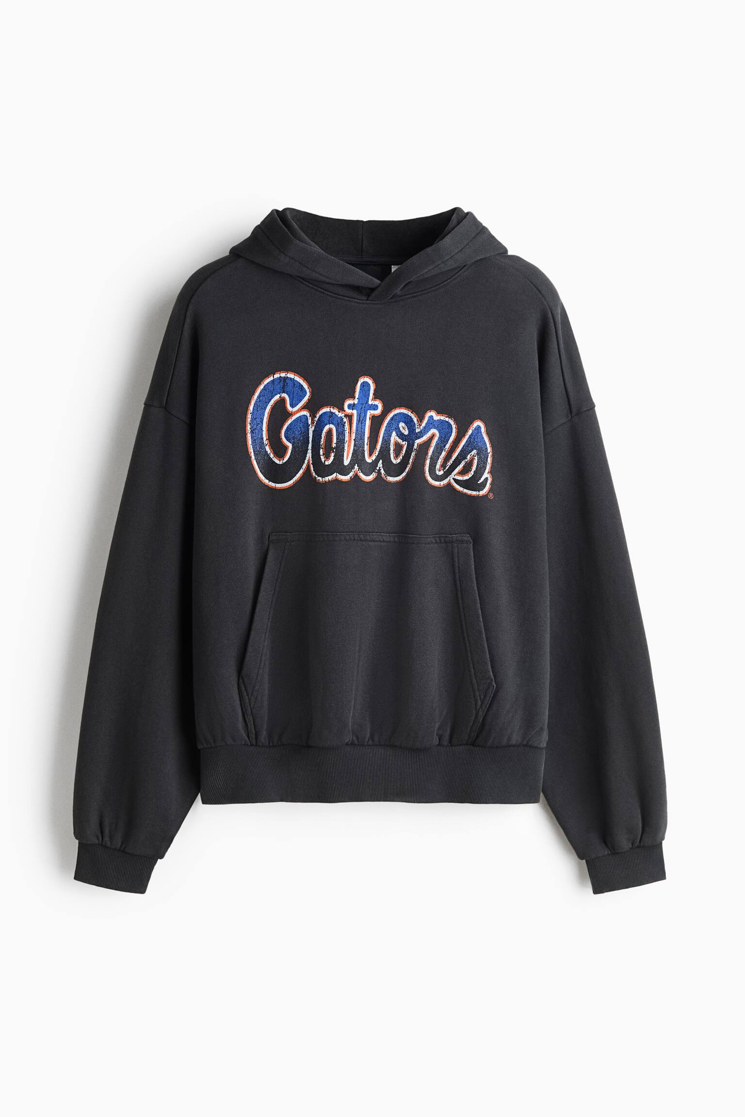 A dark grey oversized hoodie is shown from the front, featuring a distressed blue and orange "Gators" graphic printed across the chest. The design includes a wrap-style hood, dropped shoulders, a front kangaroo pocket, and ribbed cuffs and hem.