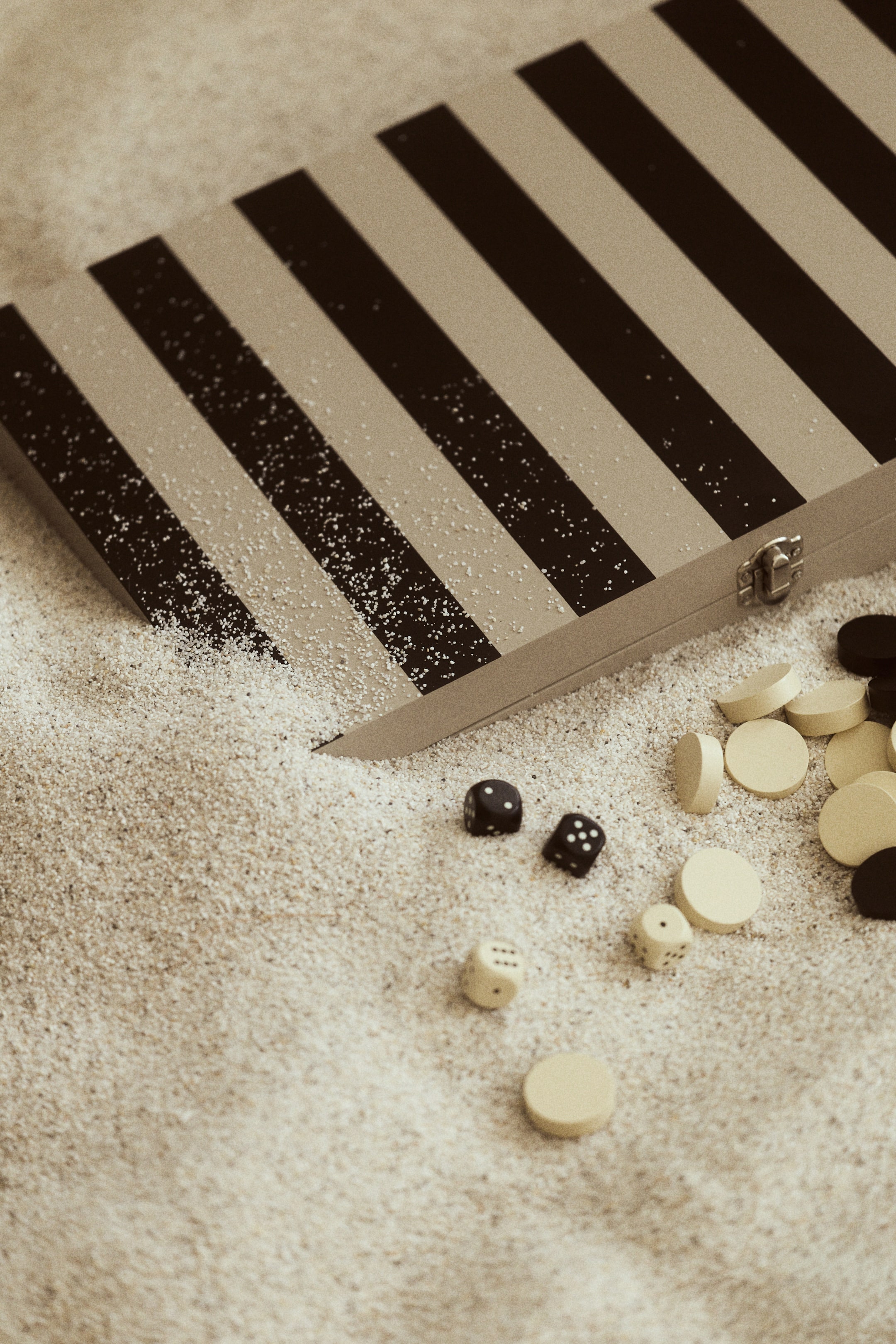 Visa större bild: Ett stängt backgammonbräde är vinklat diagonalt och visar en övre yta med växlande ljusa och mörka ränder. En metallspänne syns på sidan, och flera ljusa och mörka backgammonbrickor och tärningar är utspridda runtomkring.