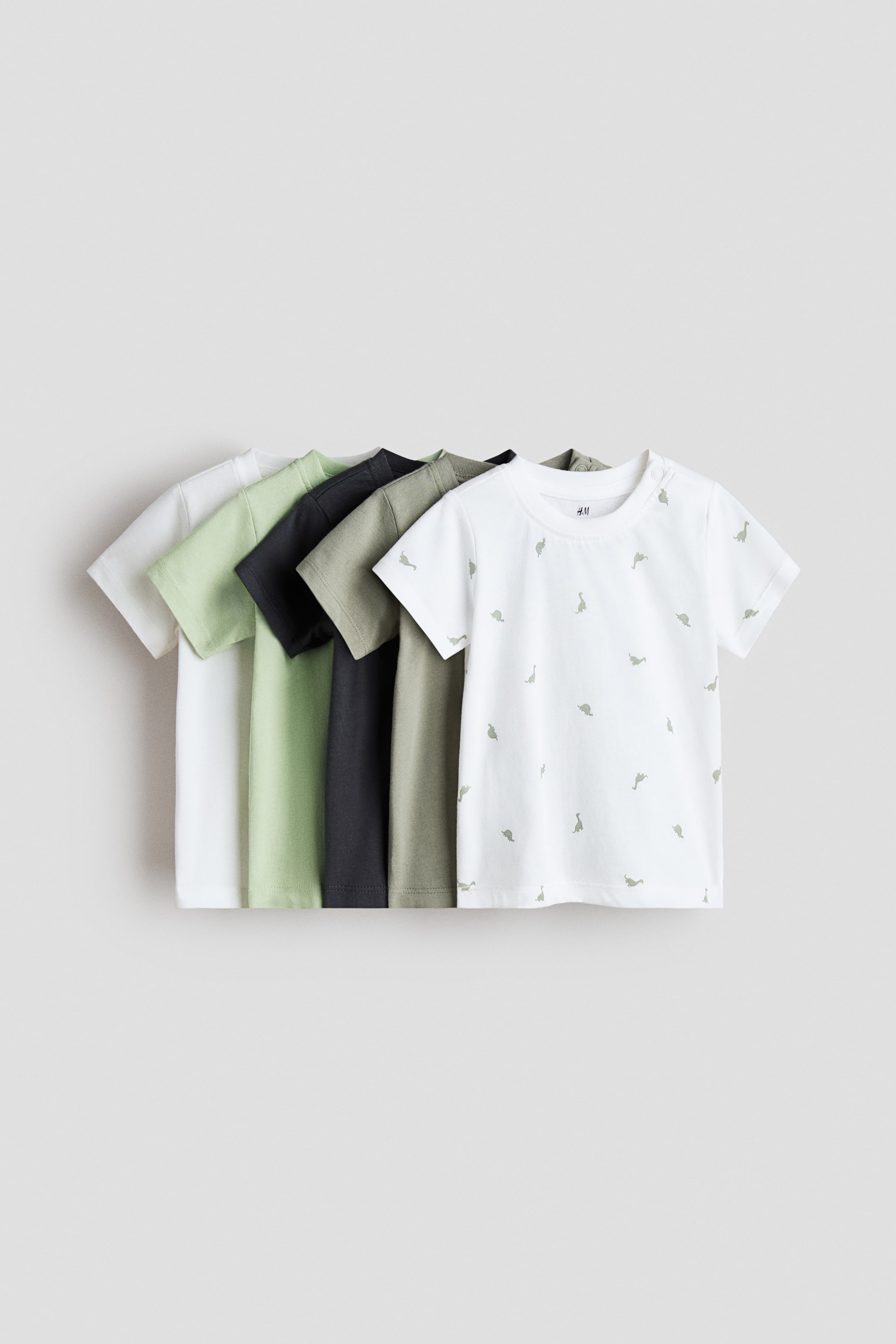 View larger image: Five short-sleeved T-shirts are shown from the front, fanned out from left to right. The white T-shirt in the foreground features a scattered light green dinosaur print, a round neck and two snap buttons on the left shoulder, while behind it solid T-shirts are visible in olive green, dark grey, light green and white.