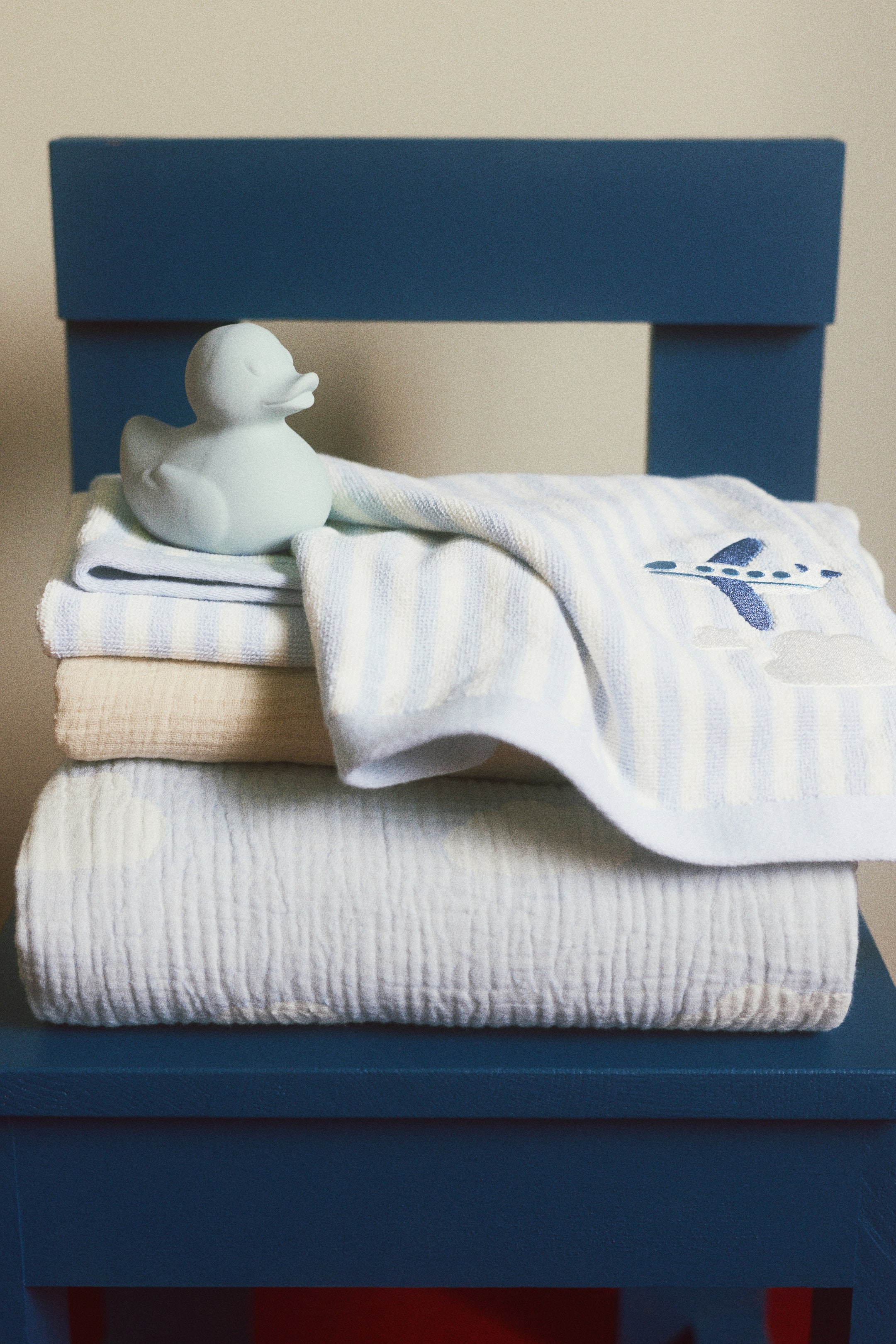 Agrandir l'image: Une pile de textiles pour bébé est soigneusement disposée, avec un canard en caoutchouc bleu clair reposant sur la serviette du dessus. La pile comprend une serviette éponge rayée bleu clair et blanc, brodée d'un avion bleu foncé et de nuages blancs, une serviette unie beige pliée, et une couverture en mousseline froissée bleu clair en bas.