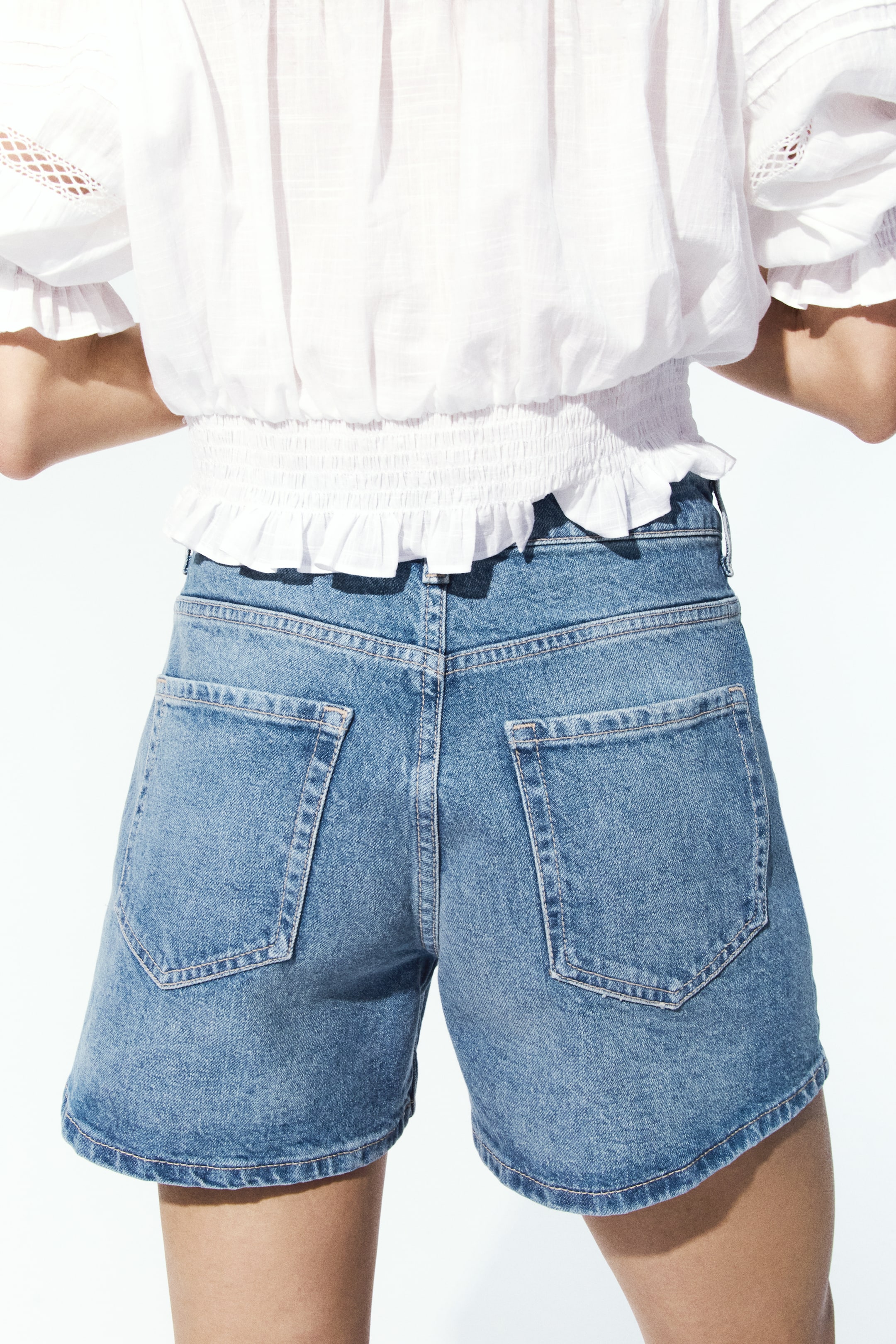 Vis større bilde: Medium blå denimshorts vises fra baksiden, med en høy midje og avslappet passform, synlig linning, beltehemper, to store skråstilte lommer og distinkte vertikale sømmer som går ned langs hvert ben og ender midt på låret.