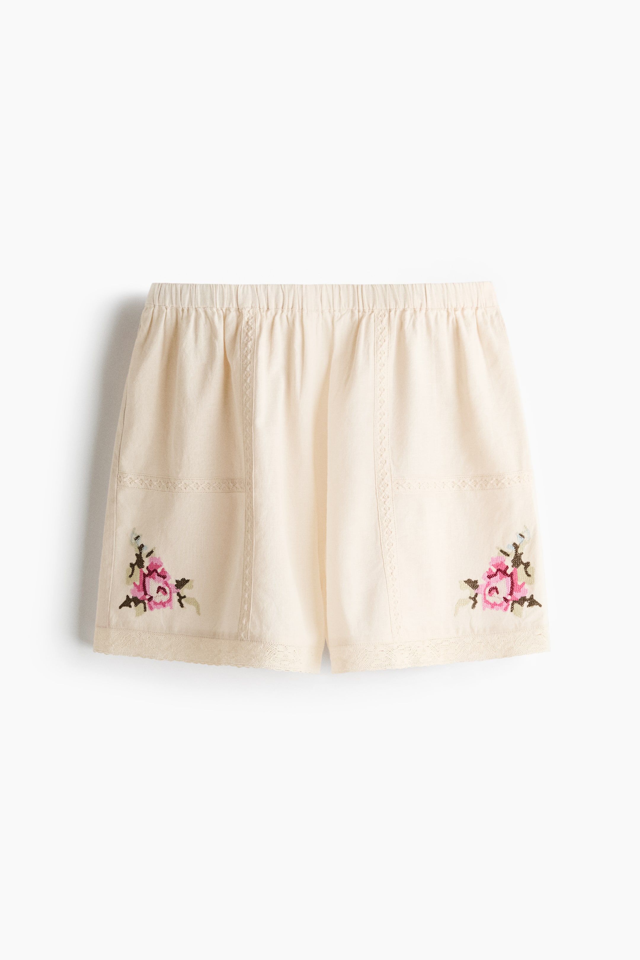 Grotere afbeelding bekijken: Crèmekleurige pyjamashorts zijn aan de voorkant gericht, met een bedekte elastische tailleband en een luchtige weving. Kantafwerkingen sieren de zoom en verticale panelen, terwijl geborduurde roze bloemmotieven verschijnen op de onderste voorkant hoeken.