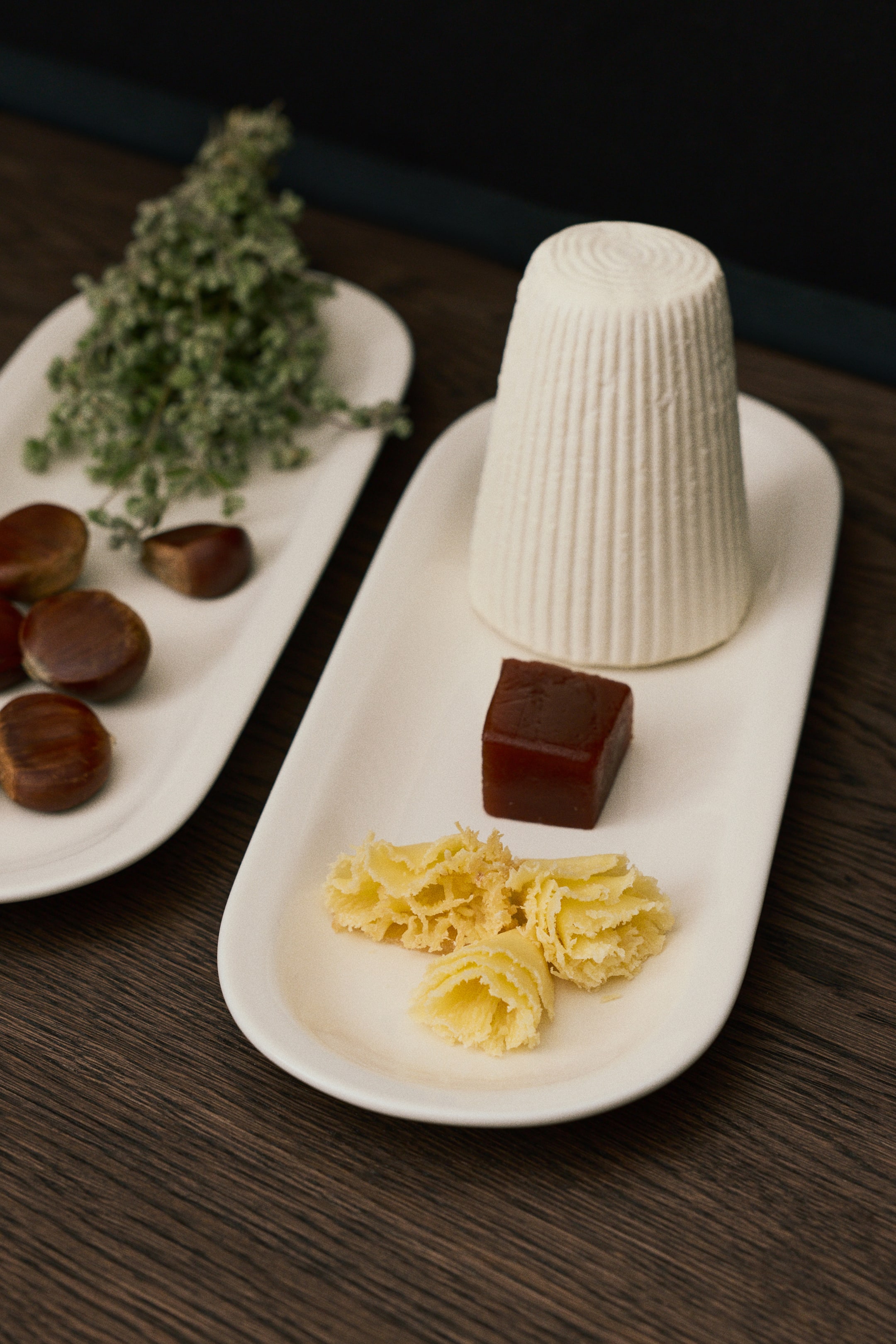 Agrandir l'image: Un plat de service ovale en porcelaine blanche présente un design minimaliste avec un bord étroit, légèrement relevé. Le plat allongé accueille sur sa surface lisse un grand fromage blanc strié, un cube de gelée sombre et des rosettes de fromage râpé, mettant en valeur sa silhouette fine et son échelle fonctionnelle.