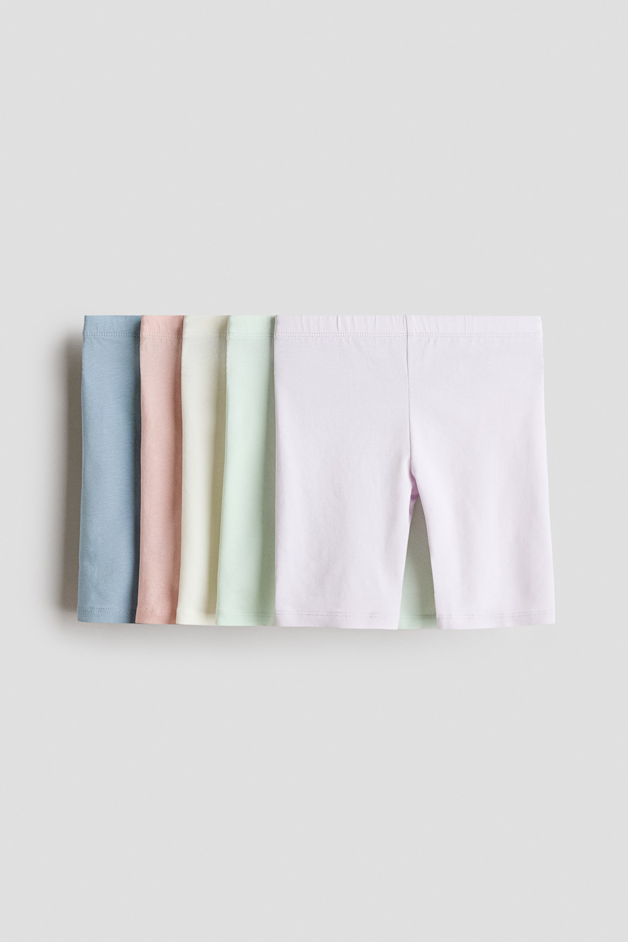 Größeres Bild anzeigen: Radlerhosen aus weichem Baumwolljersey mit elastischem Bund werden in einem Set von fünf Pastellfarben präsentiert. Jedes Paar ist leicht überlappend, wobei die Vorderseite der weißen Shorts rechts zu sehen ist, während andere Paare in Blau, Rosa, Creme und Hellgrün dahinter sichtbar sind.