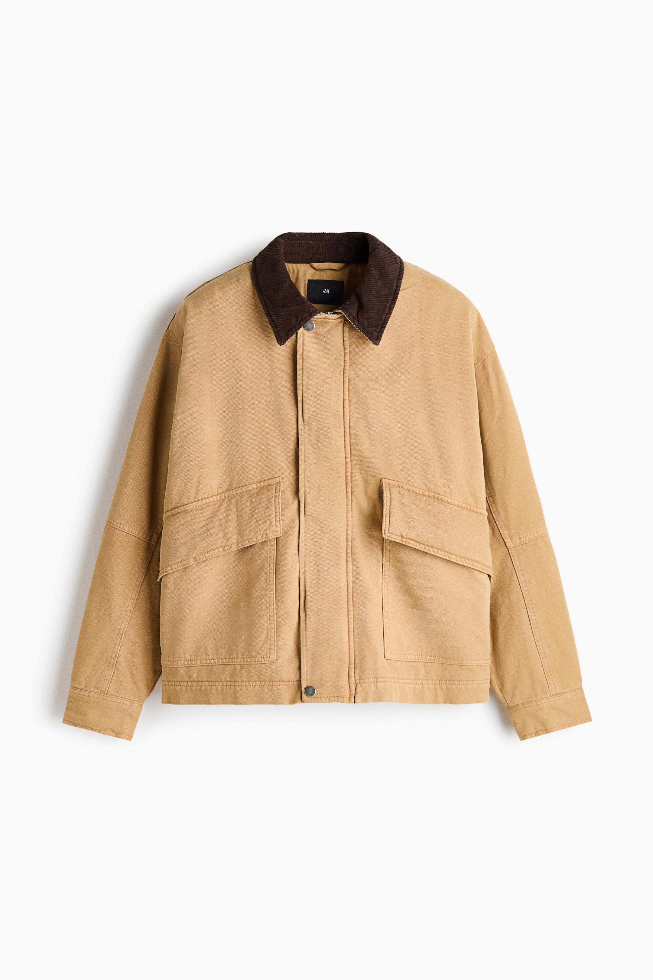 View larger image: A tan canvas utility jacket is shown from the front, featuring a contrasting dark brown corduroy collar and a concealed front closure with visible snap buttons at the top and bottom. The jacket has two large front flap pockets and long sleeves with panel seam detailing.