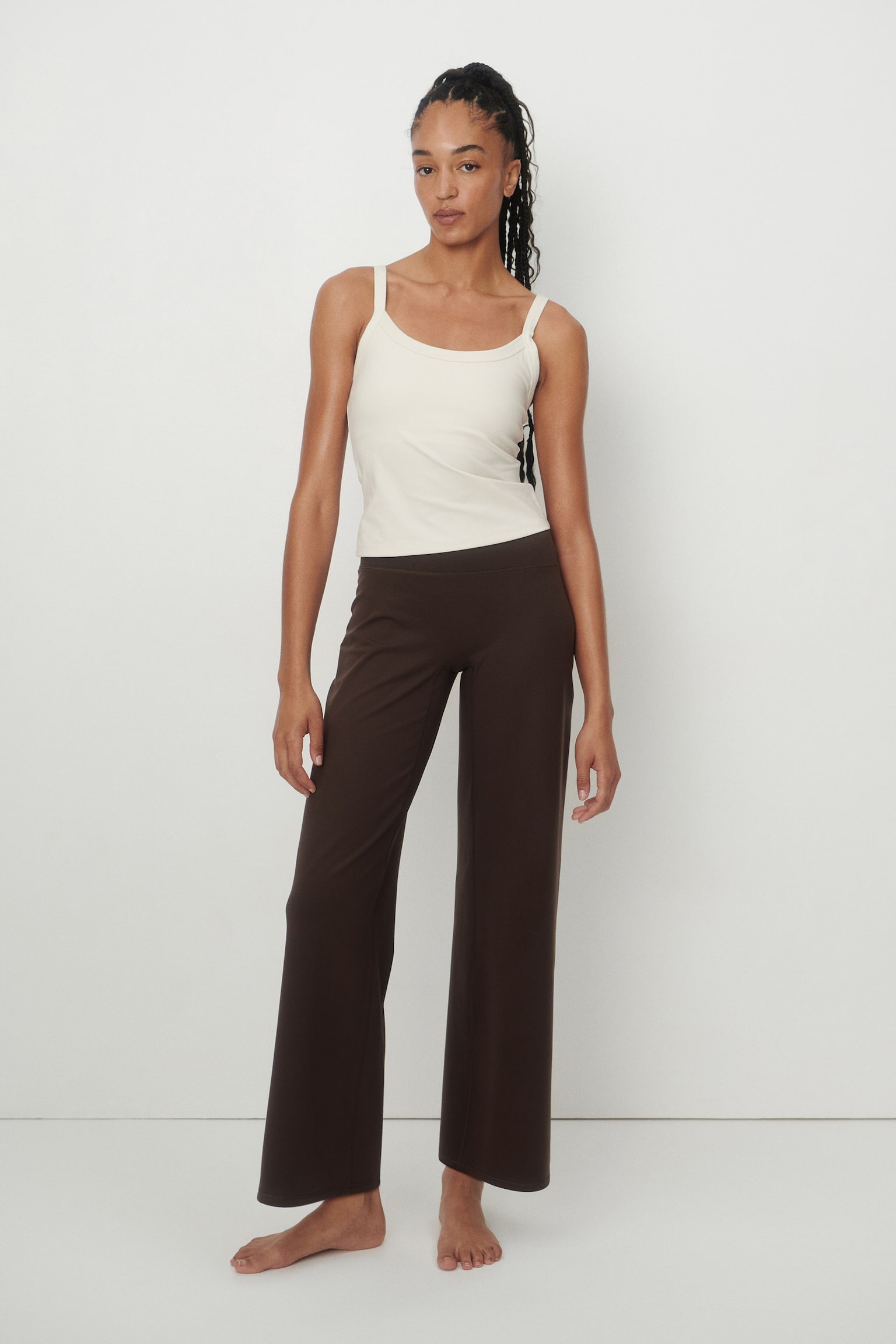 The model is wearing an integrated-bra sports vest top. The cream-coloured top features spaghetti straps and subtle vertical ribbing, styled with dark brown wide-leg trousers. - 1