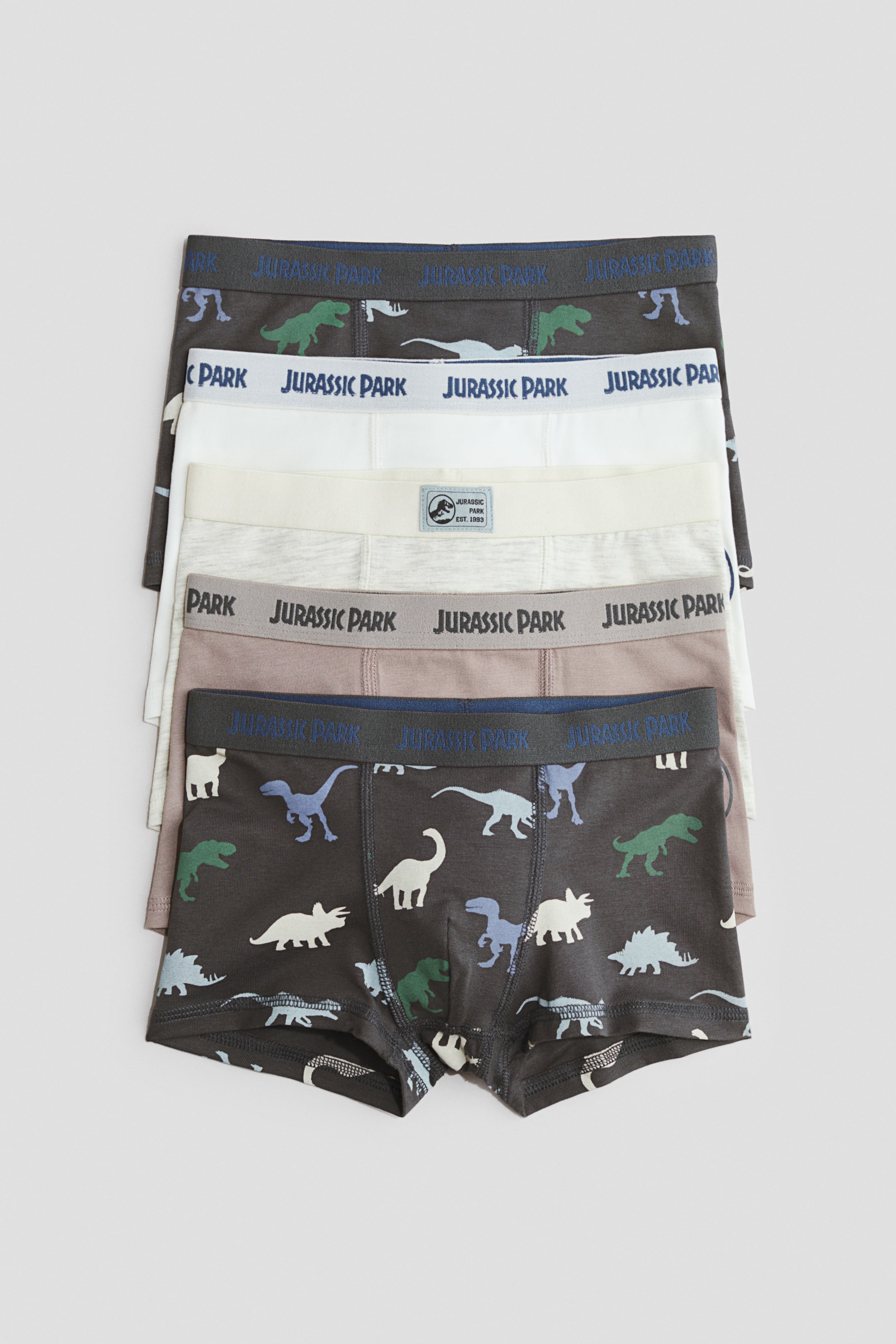 Zobacz większy obraz: Pięciopak bawełnianych bokserek typu boxer briefs jest ułożony pionowo, ukazując ich przednie wzory z krótkimi nogawkami i elastycznymi pasami. Zestaw obejmuje dwie pary w ciemnoszarym kolorze z wielobarwnym nadrukiem dinozaurów oraz trzy pary w jednolitych kolorach: białym, jasnoszarym i taupe, wszystkie z brandingiem Jurassic Park na pasach.
