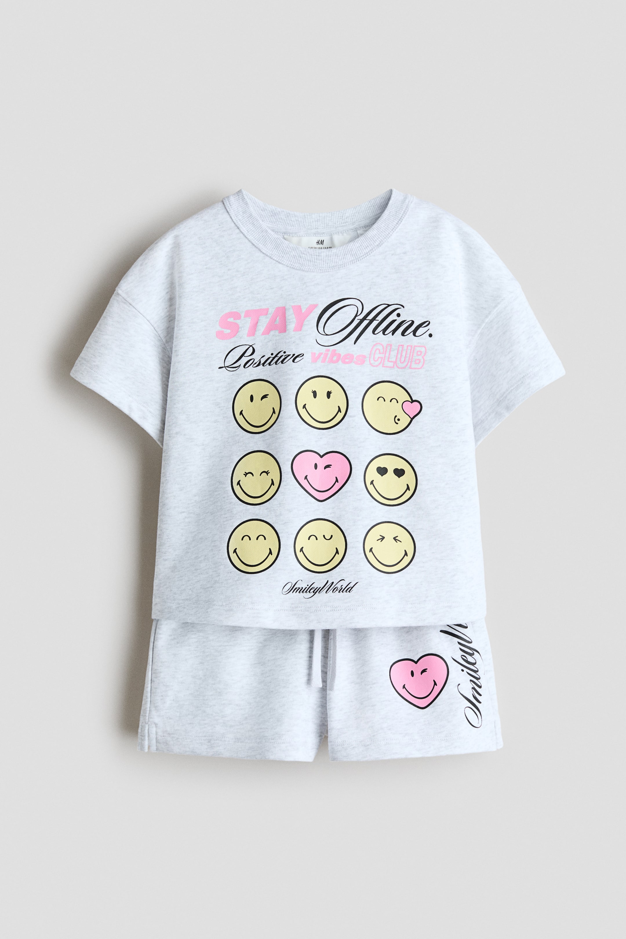 View larger image: A light gray sweatshirt fabric set is showcased from the front, consisting of a T-shirt and shorts. The T-shirt features a ribbed neckline, dropped shoulders, and a front print with the text 'STAY Offline. Positive vibes CLUB' alongside a grid of nine emoji-like faces, including eight yellow smileys and one pink heart-shaped smiley. The shorts have an elasticated drawstring waist and a pink heart-shaped smiley print on the left leg.