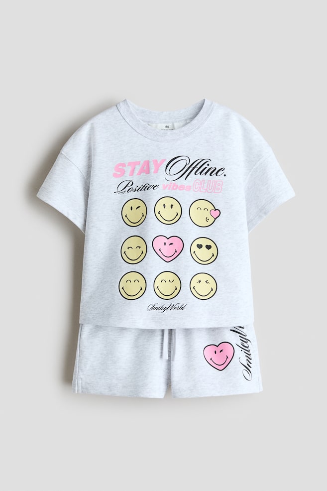 Un ensemble en tissu molleton gris clair est présenté de face, comprenant un T-shirt et un short. Le T-shirt arbore un col à bord-côtes, des épaules tombantes et un imprimé sur le devant avec le texte "STAY Offline. Positive vibes CLUB" et une grille de neuf visages ressemblant à des émojis, incluant huit smileys jaunes et un smiley en forme de cœur rose, tandis que le short présente une taille élastiquée avec cordon de serrage et un imprimé smiley en forme de cœur rose sur la jambe gauche. - 1