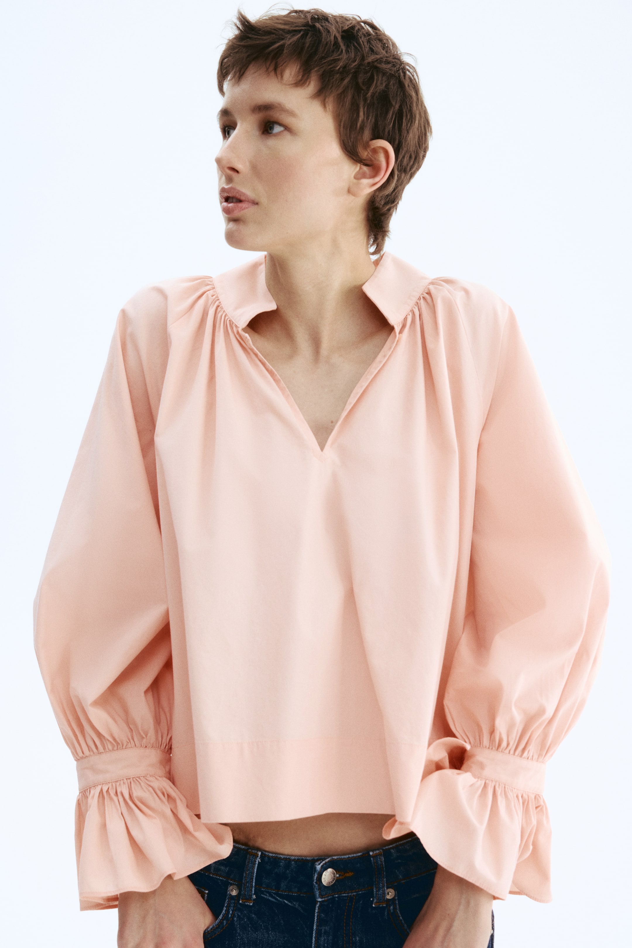 Nagyobb kép megtekintése: Egy világos rózsaszín blúz látható elölről-jobbról, állógallérral és mély V-kivágással. A vállaknál ráncolt részletekkel tervezték, amelyek bő, hosszú ujjakhoz vezetnek, amelyek fodros mandzsettával végződnek.