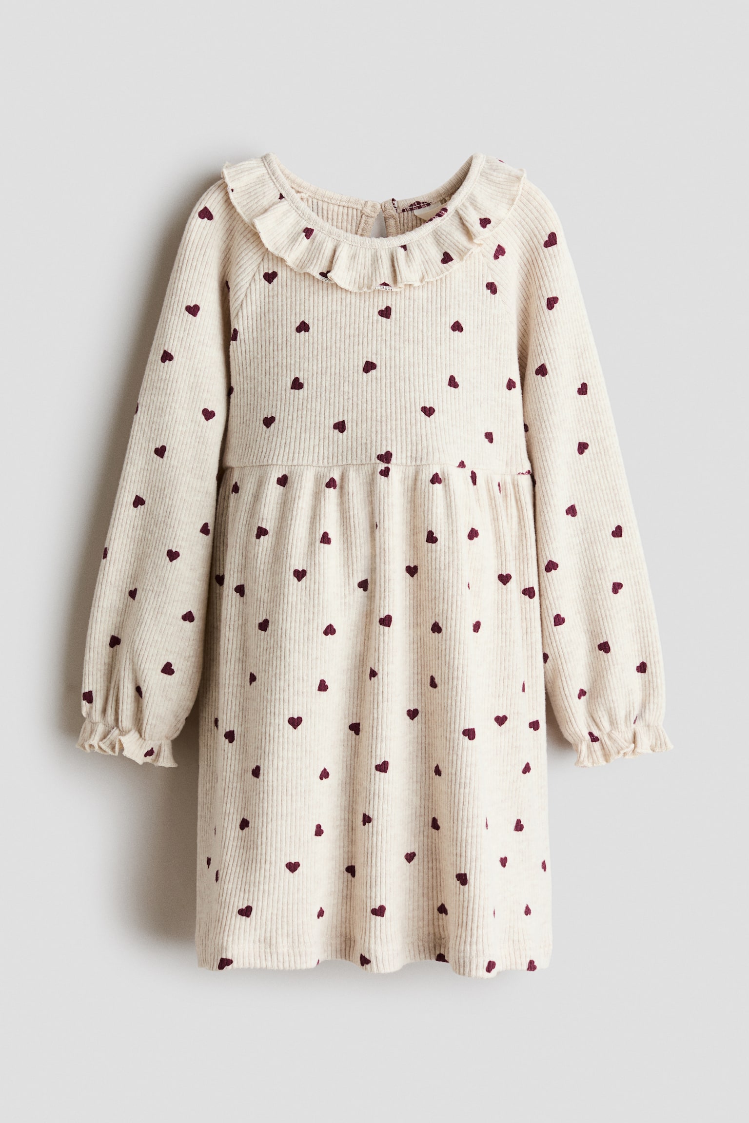 Ein cremefarbenes, geripptes Jerseykleid mit einem durchgehenden Muster aus kleinen roten Herzen ist von vorne zu sehen. Das Kleid zeigt einen runden, mit Rüschen besetzten Ausschnitt, lange Ãrmel mit gerüschten Abschlüssen und eine Taillennaht mit Kräuselung, die in einen sanft ausgestellten Rock übergeht.