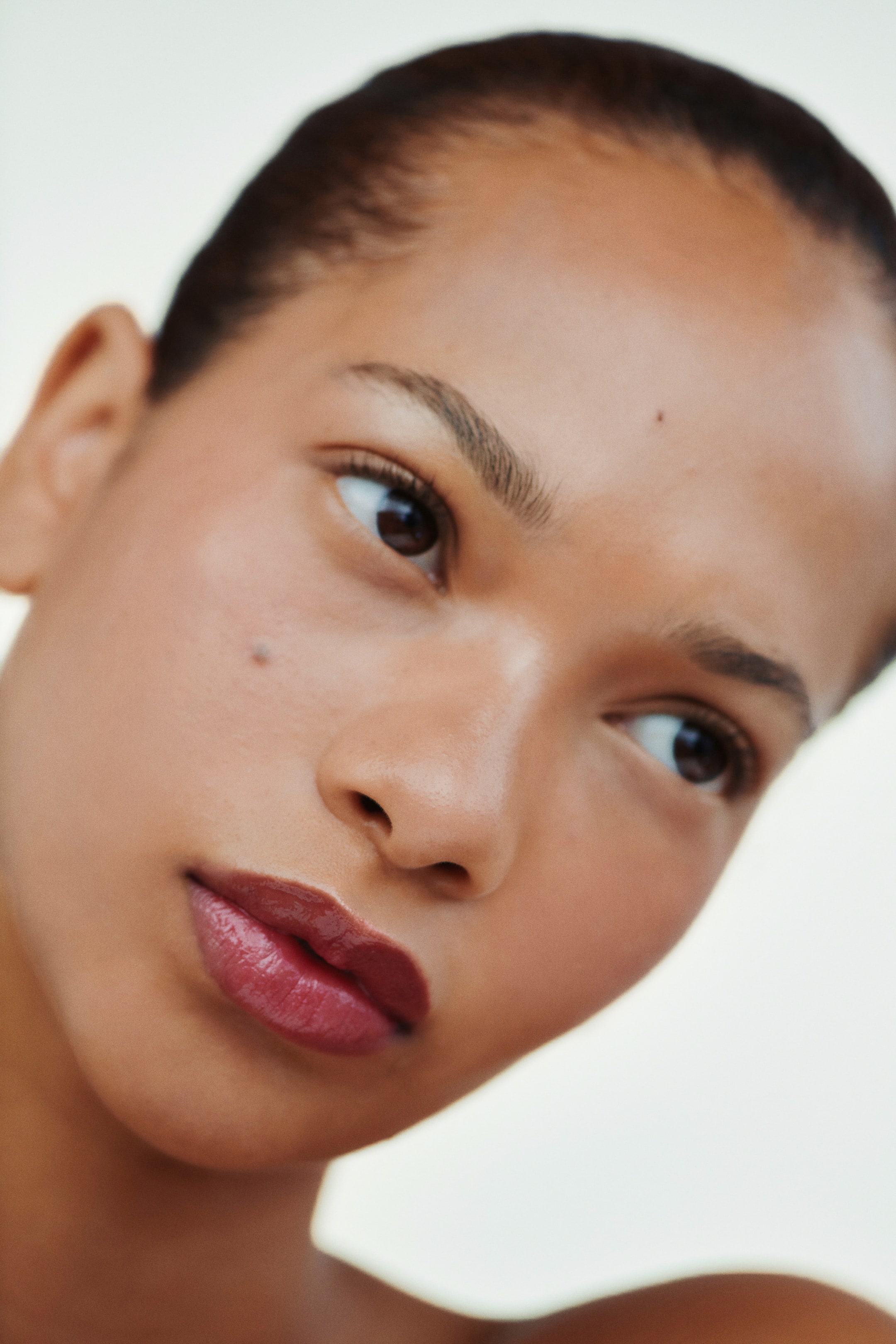 Größeres Bild ansehen: Tiefes Beeren-Lipgloss bedeckt die Lippen und sorgt für ein hochglänzendes, volles Finish. Die Haut zeigt einen glatten, taufrischen Teint mit subtilen Highlights auf den Wangenknochen und dem Nasenrücken, alles aus einem leicht erhöhten Blickwinkel betrachtet.