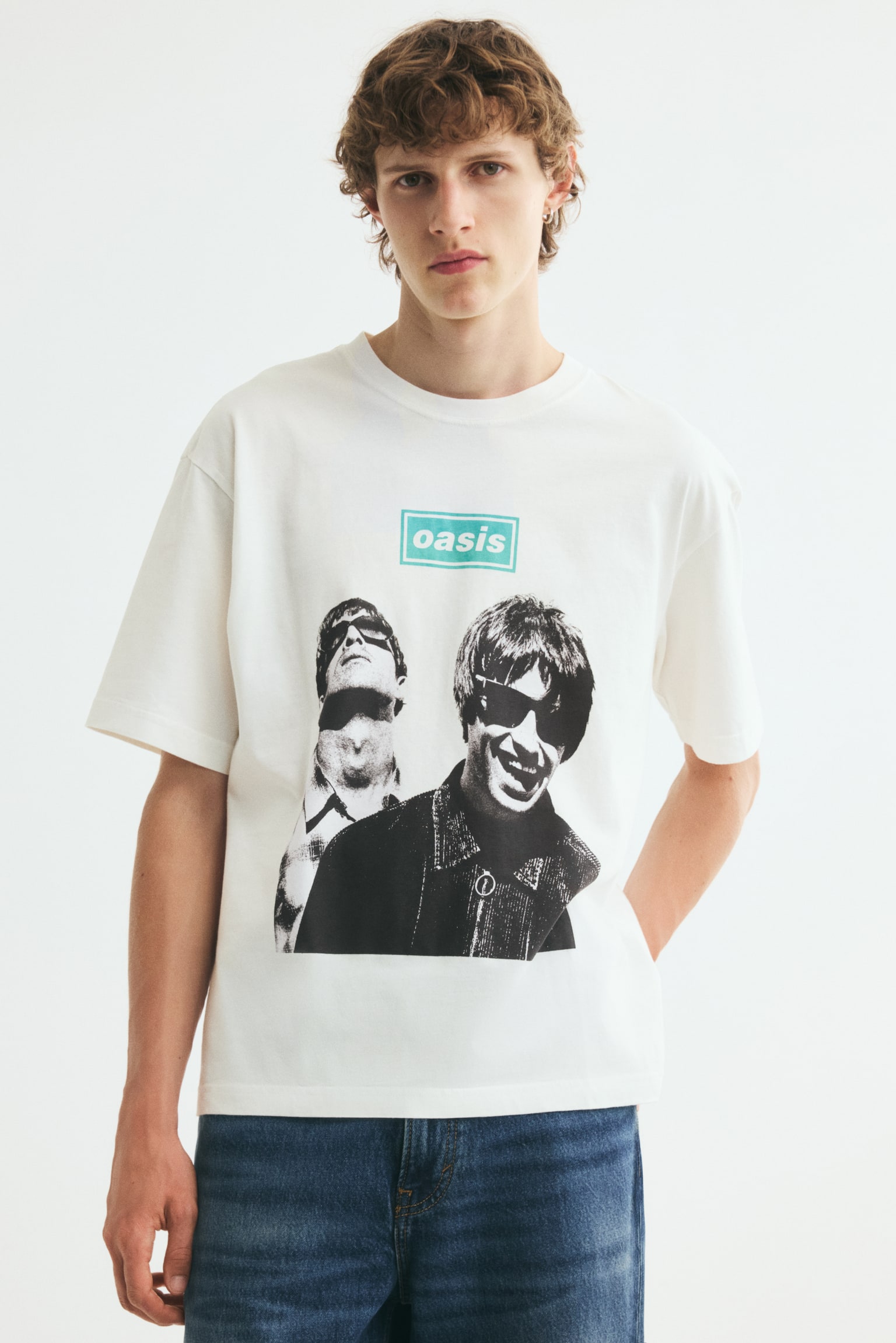 A cream-colored, relaxed-fit T-shirt is being worn by the model, featuring a ribbed round neck, dropped shoulders, and a black-and-white graphic print of the band Oasis with a teal logo on the front. The T-shirt is styled with blue denim jeans, with the model standing and resting one hand behind their hip. - 1
