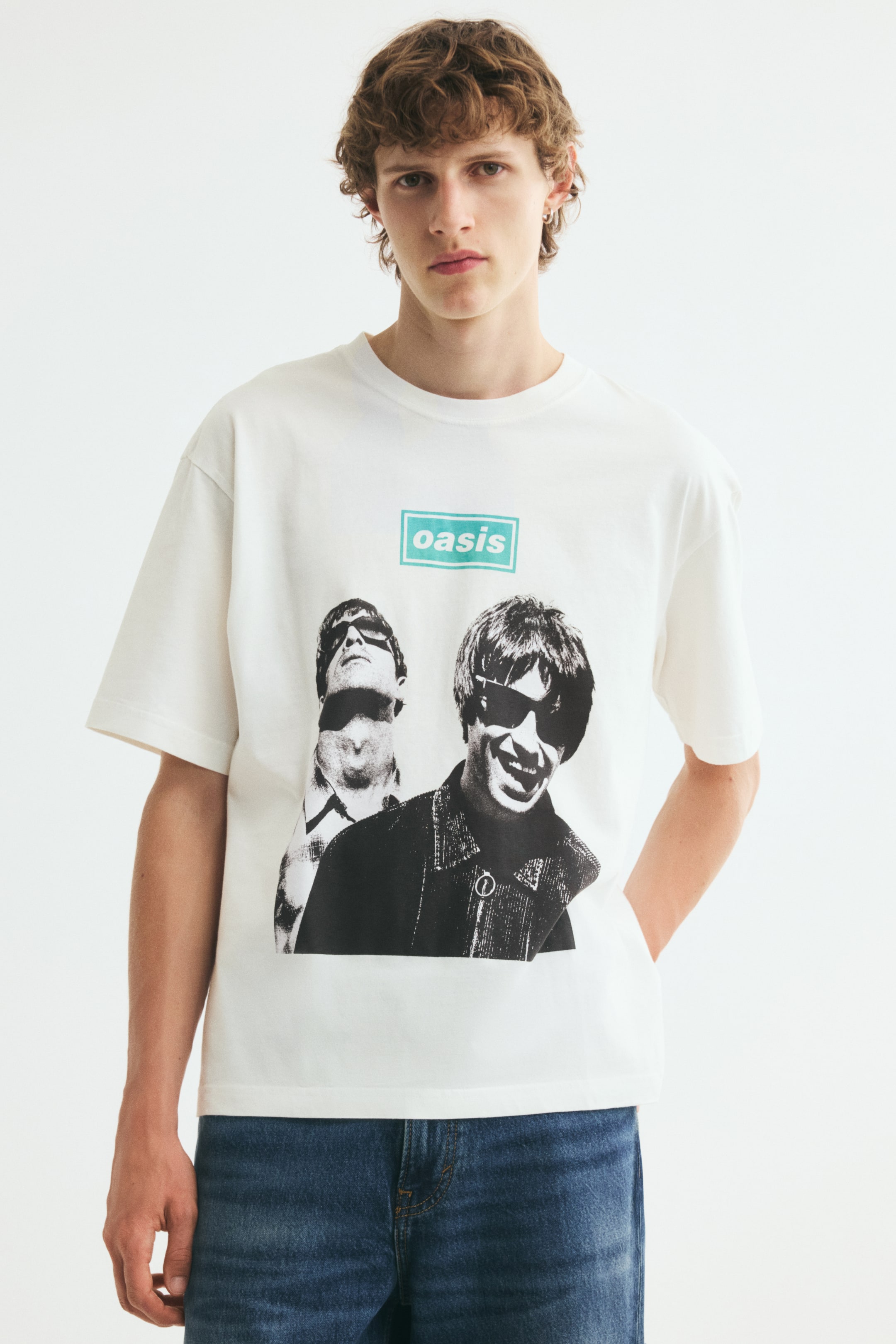 Ver imagen más grande: Una camiseta de corte relajado en color crema la lleva puesta la modelo, con cuello redondo acanalado, hombros caídos y un estampado gráfico en blanco y negro de la banda Oasis con un logotipo en color turquesa en la parte frontal. La camiseta se combina con unos vaqueros de denim azul, y la modelo aparece de pie, apoyando una mano detrás de la cadera.