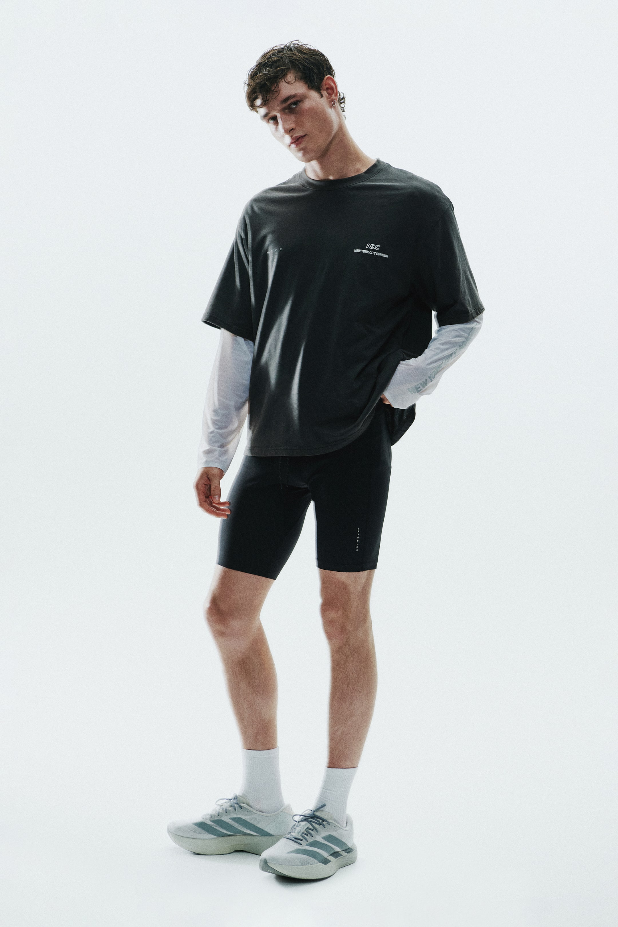 View larger image: A black short-sleeved T-shirt featuring a small white chest graphic is worn by the model over a white long-sleeved top and paired with black athletic compression shorts. This layered look is styled with white crew socks and light grey running shoes.