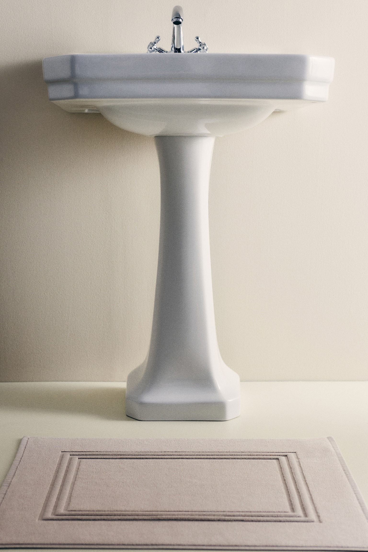 A rectangular bath mat in a light beige cotton terry is presenting its front. It is featuring a subtle sheen and a raised, rectangular frame detailing its center, with a woven trim finishing its outer edges. - 1