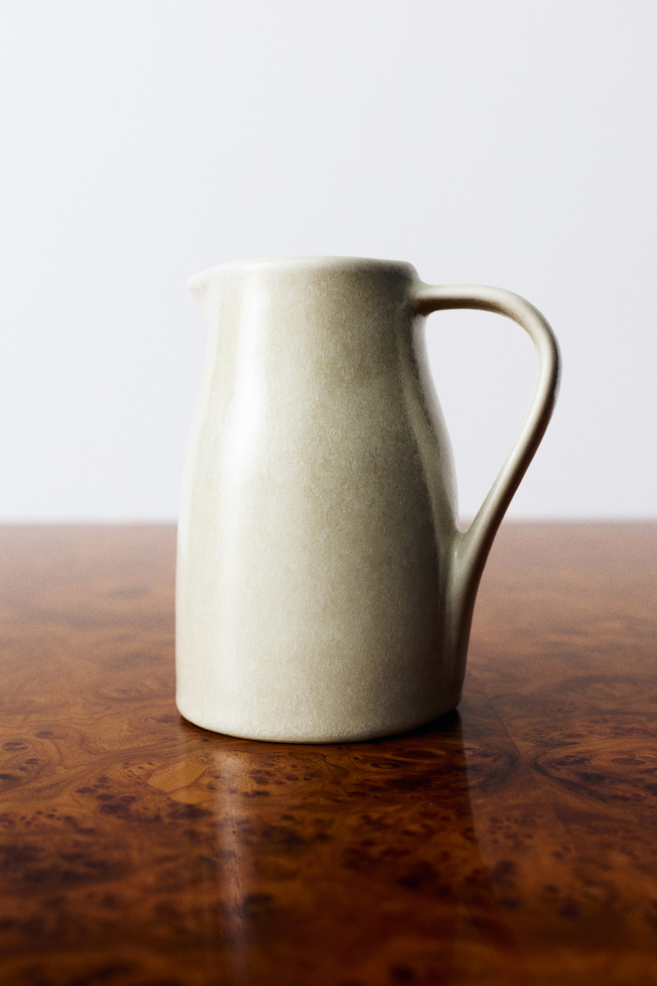 Visualizza immagine più grande: Una brocca per latte in gres con corpo rastremato e smalto reattivo punteggiato color beige chiaro è mostrata in profilo laterale. La brocca presenta un piccolo beccuccio versatore integrato su un lato e un sottile manico ad anello ricurvo sull’altro.