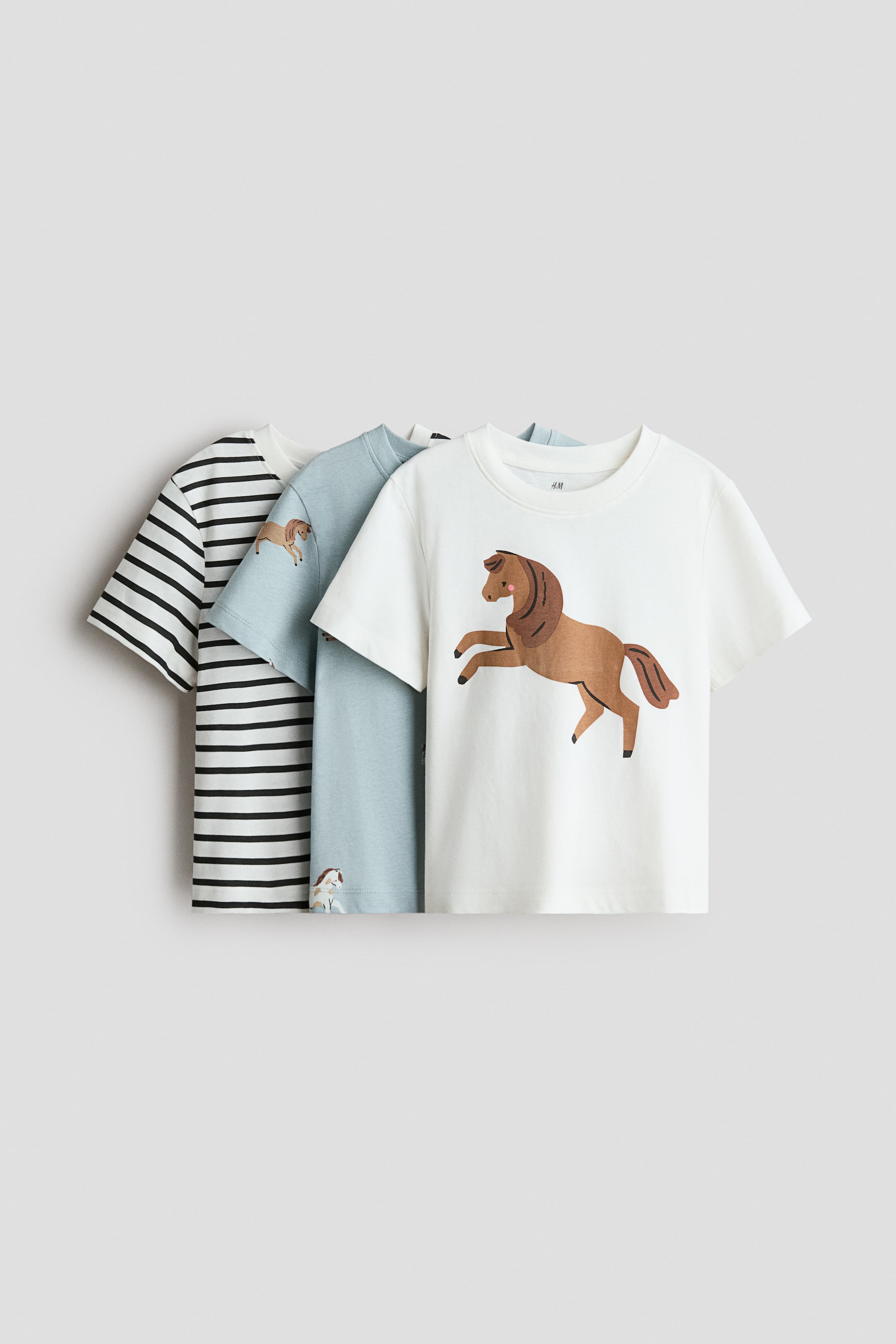 View larger image: Three cotton jersey T-shirts are shown in a front-facing view in an overlapping arrangement, each featuring a round rib-trimmed neckline and short sleeves. The front white T-shirt features a large graphic of a rearing brown horse, while the middle light blue T-shirt and the back black-and-white striped T-shirt are layered behind it.