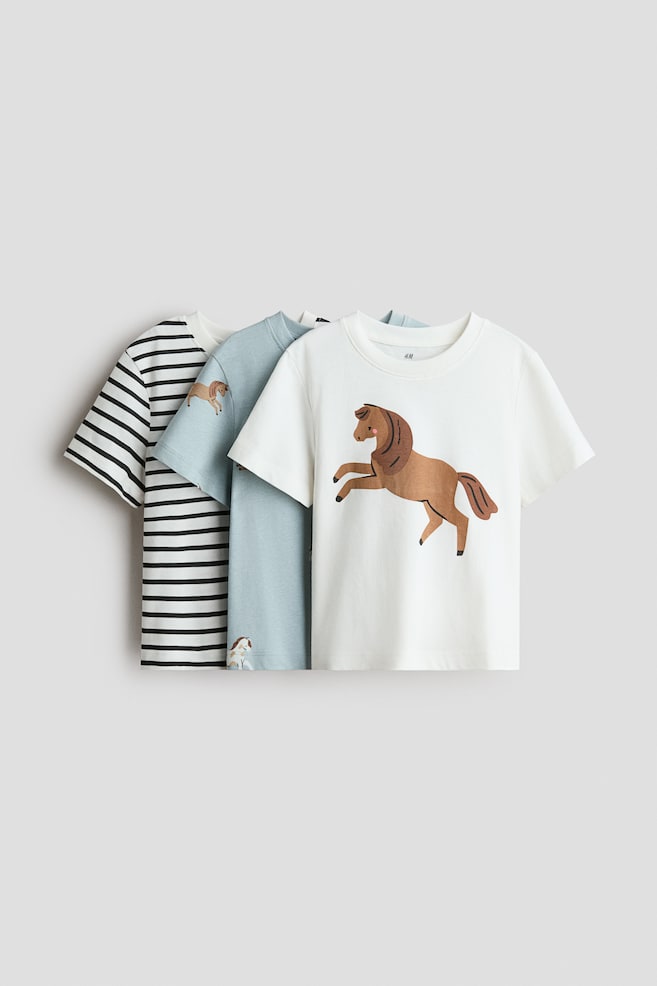Three cotton jersey t-shirts are displaying a front-facing view in an overlapping arrangement, each featuring a round, rib-trimmed neckline and short sleeves. The front-most white tee is featuring a large graphic of a rearing brown horse, while the middle light blue tee and back-most black-and-white striped tee are layering behind it. - 1