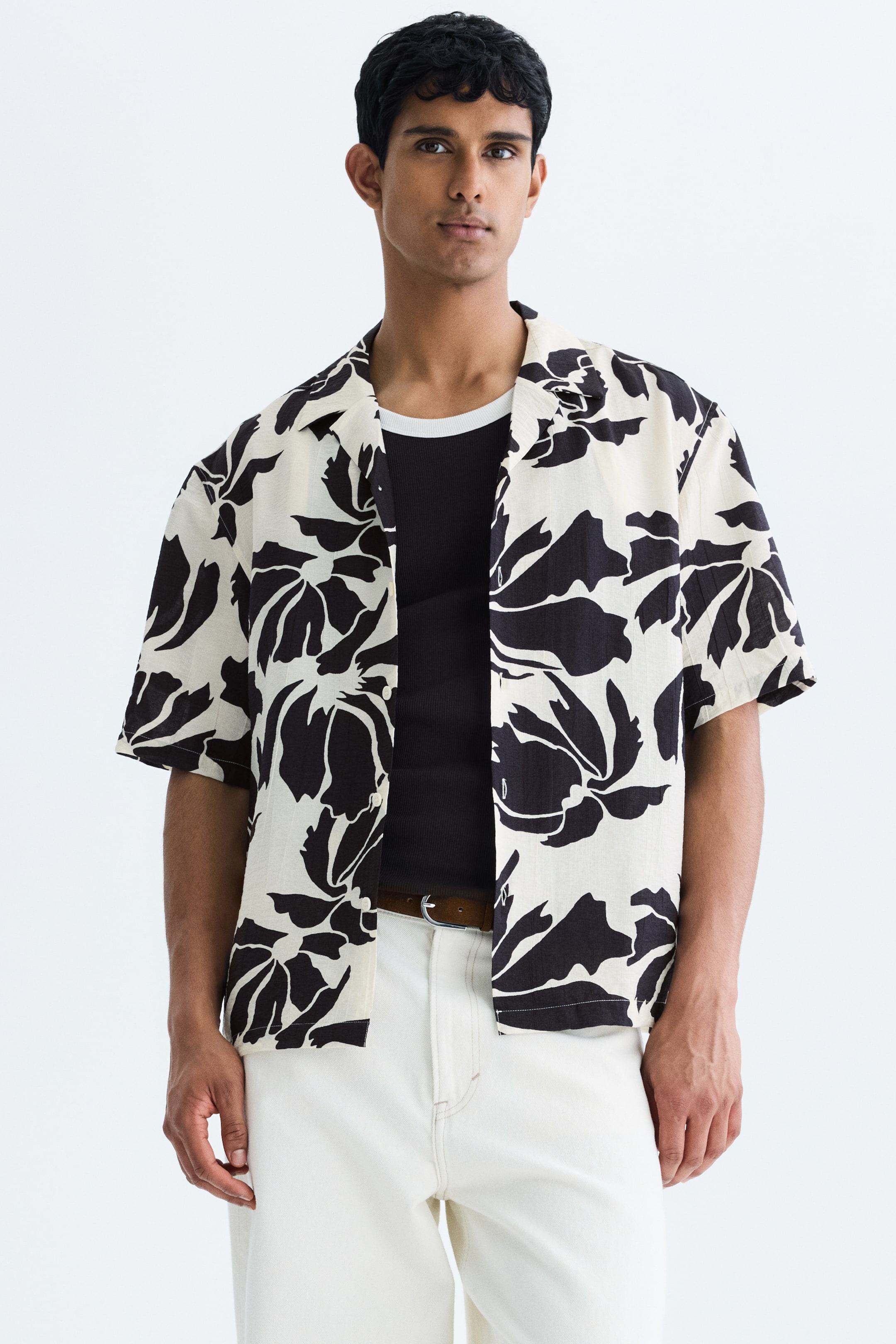View larger image: The model is wearing a short-sleeved shirt featuring a black and white floral pattern and a resort collar. This relaxed-fit shirt, with its straight-cut hem, is styled open over a black tank top and paired with white trousers and a brown belt.