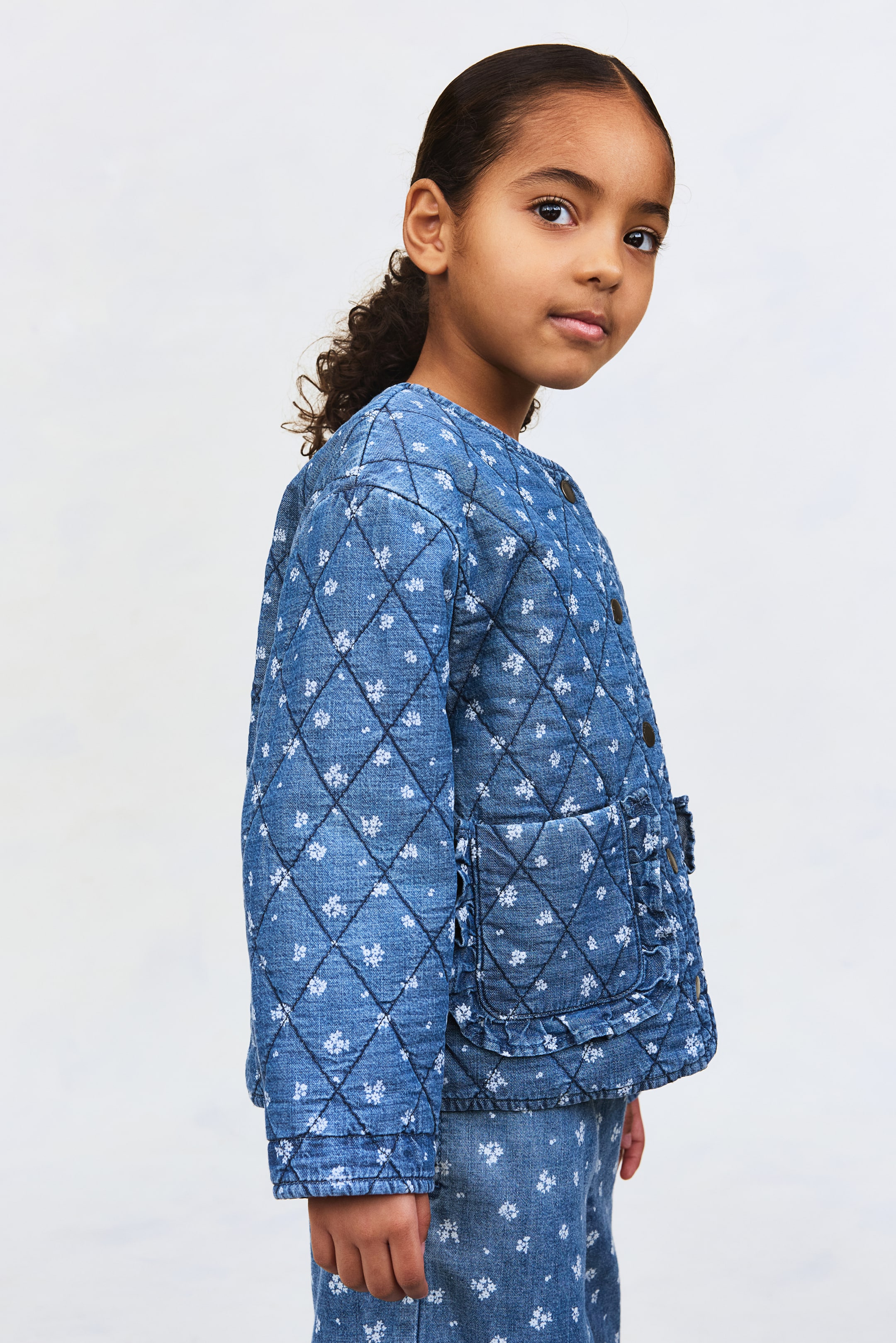 Visa större bild: En blå quiltad denimjacka med ett vitt blommönster och diamantkviltning presenteras från en lätt sidovinkel, med rund halsringning, knäppning framtill och påsydda fickor med volangkant. Matchande blå denimbyxor med samma blommönster syns.