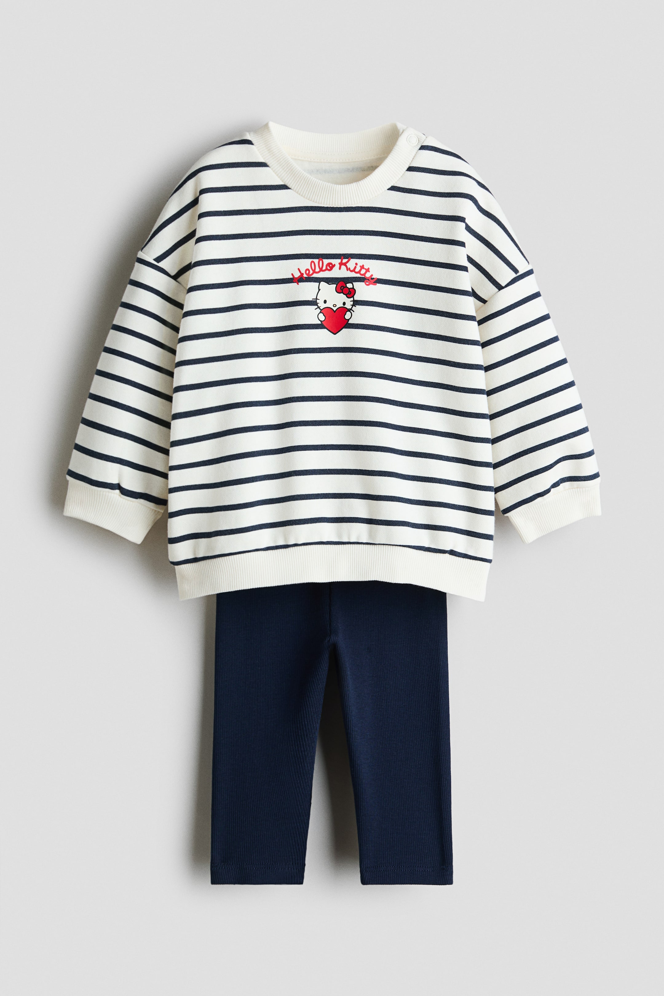 Grotere afbeelding bekijken: Een 2-delig sweatshirt- en legging-set bestaat uit een vooraanzicht gestreepte sweatshirt en effen leggings. De gebroken witte sweatshirt heeft navyblauwe horizontale strepen, een geribde ronde hals, manchetten en zoom, samen met een geborduurd "Hello Kitty"-logo met een karakter dat een rood hart vasthoudt op de borst en twee witte drukknoopjes op de linkerschouder. De navyblauwe leggings hebben een geribde textuur en een nauwsluitende pasvorm.