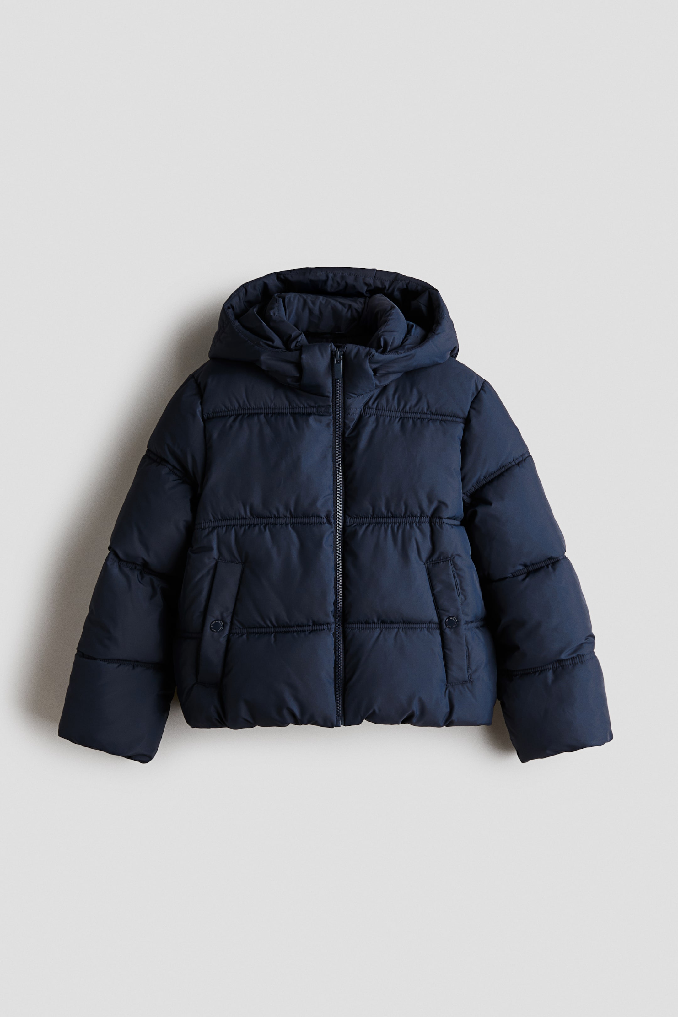 Ampliar la imagen: Una chaqueta puffer azul marino está de frente, mostrando un diseño acolchado horizontal, un cuello alto tipo chimenea con capucha integrada y un cierre central con cremallera. La chaqueta cuenta con dos bolsillos laterales de ojal en la parte frontal con cierres a presión y mangas largas.