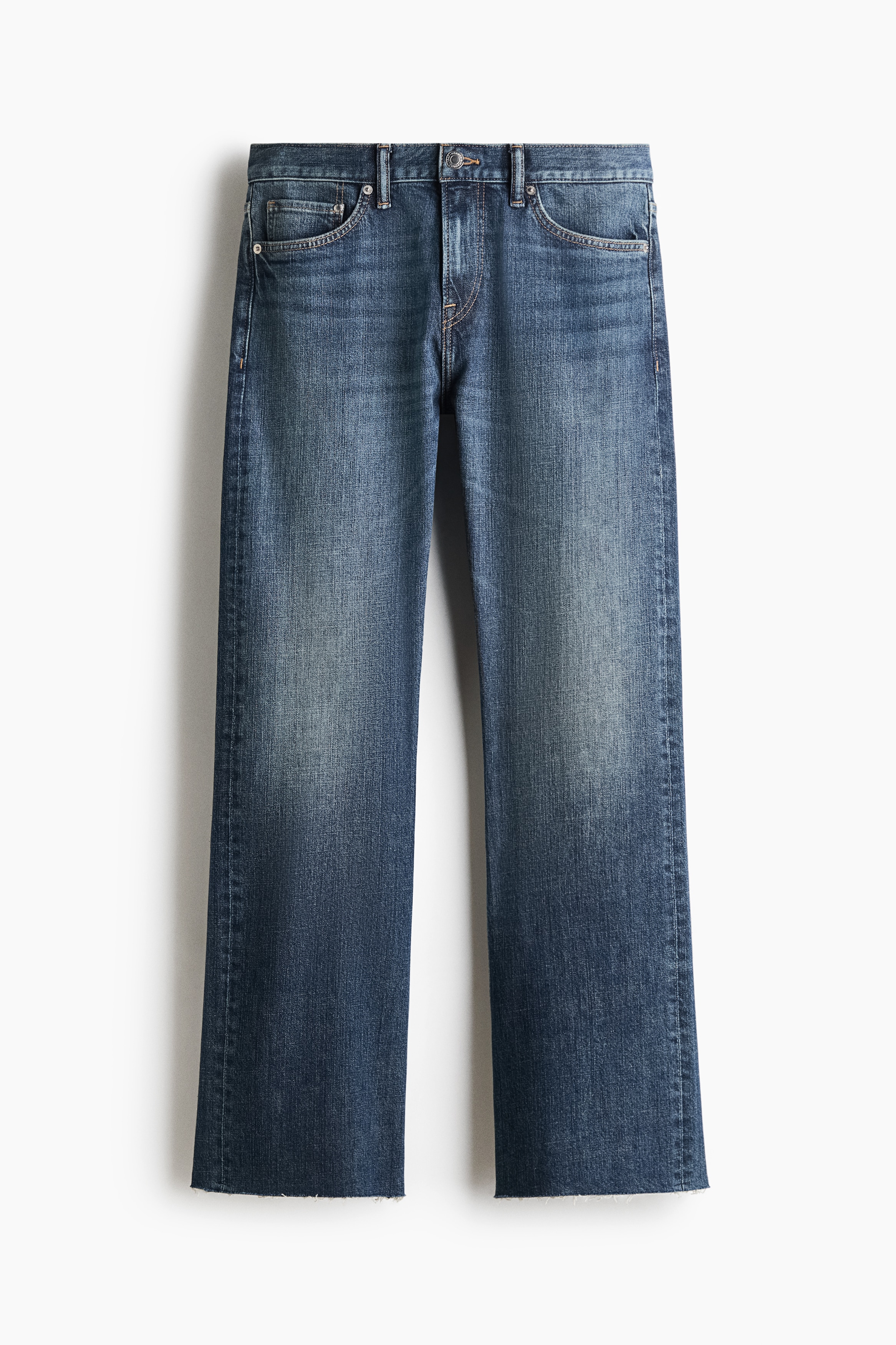 Et par blå denimjeans i medium-vask med lige ben bæres af modellen og har rå kant forneden samt et klassisk design med fem lommer. Jeansene styles med et sort læderbælte, en lyseblå skjorte med knapper lagt over en hvid T-shirt og sorte lædersko.