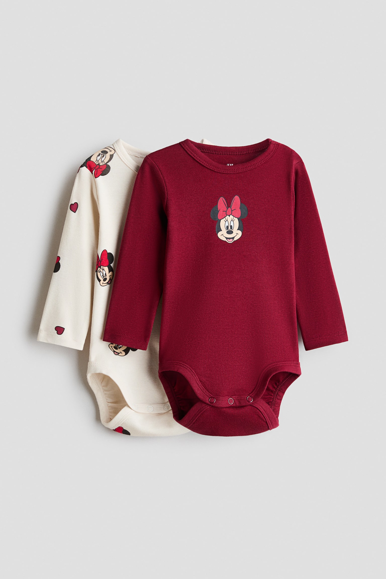 Dva bebi bodija dugih rukava su preklopljena i okrenuta ka prednjoj strani. Prednji bodi je u jednobojnoj tamnocrvenoj boji sa malim grafiÄkim prikazom Minnie Mouse na grudima, dok je drugi bodi u prljavo beloj boji sa sveobuhvatnim printom Minnie Mouse i malih crvenih srca.