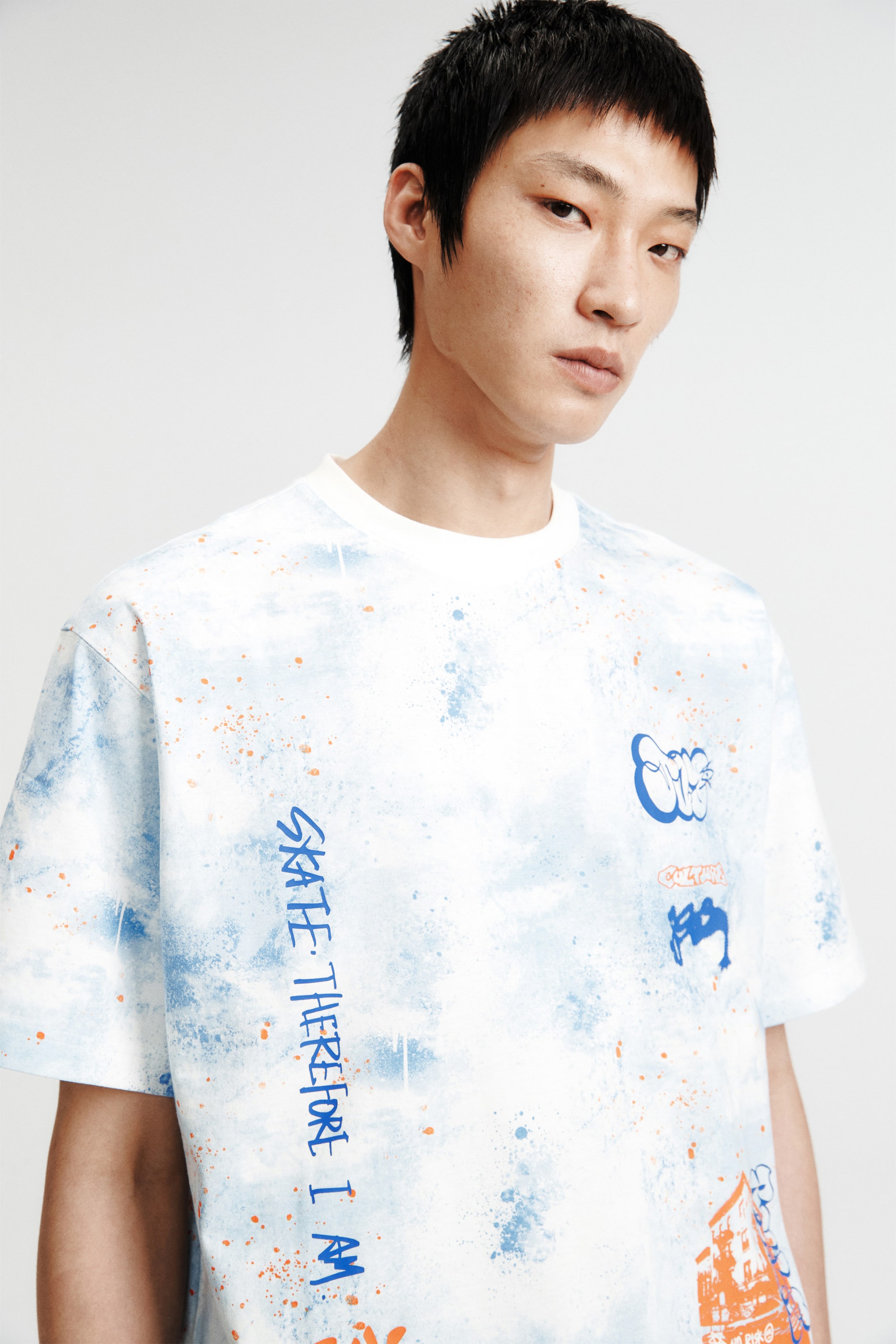 拡大画像を表示: ヘビーウェイトのプリントコットンジャージーTシャツは、ラウンドネックのリブトリム、ドロップショルダー、全体にライトブルーとホワイトのスプラッタープリントにオレンジの斑点が特徴です。モデルはルーズフィットのTシャツを着用しており、左側に縦に「SKATE. THEREFORE I AM」という青いテキストグラフィックが表示され、右胸には追加の青とオレンジのグラフィックがあります。