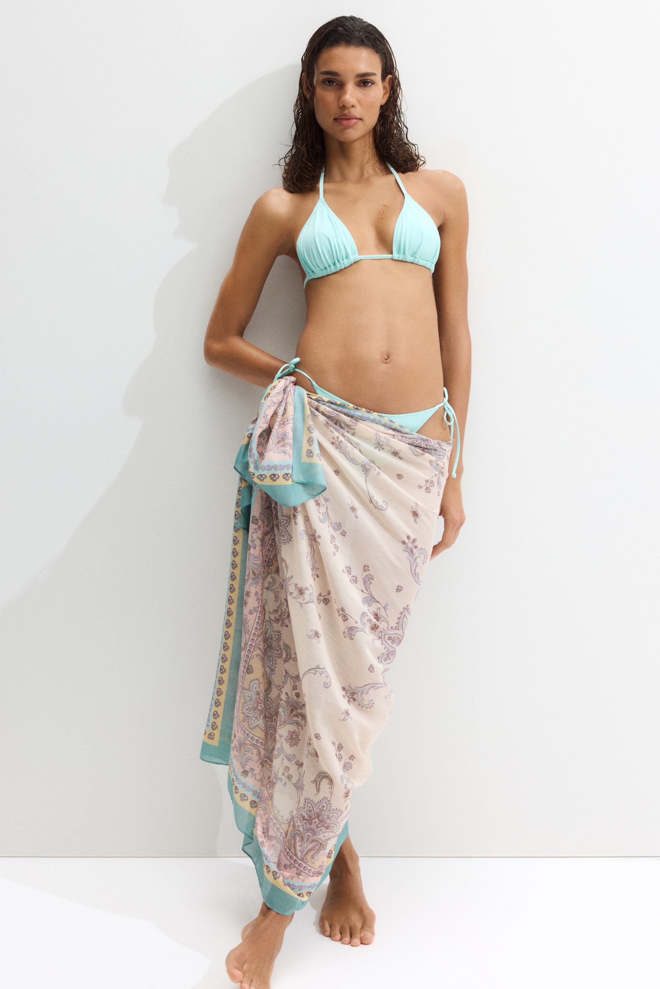 Größeres Bild ansehen: Ein Sarong in einem luftigen Gewebe, gemustert mit Paisley-Designs und einem türkisfarbenen Rand, wird vorne gebunden und als Rock getragen. Das Model trägt den Sarong über einem hellblauen Bikini.