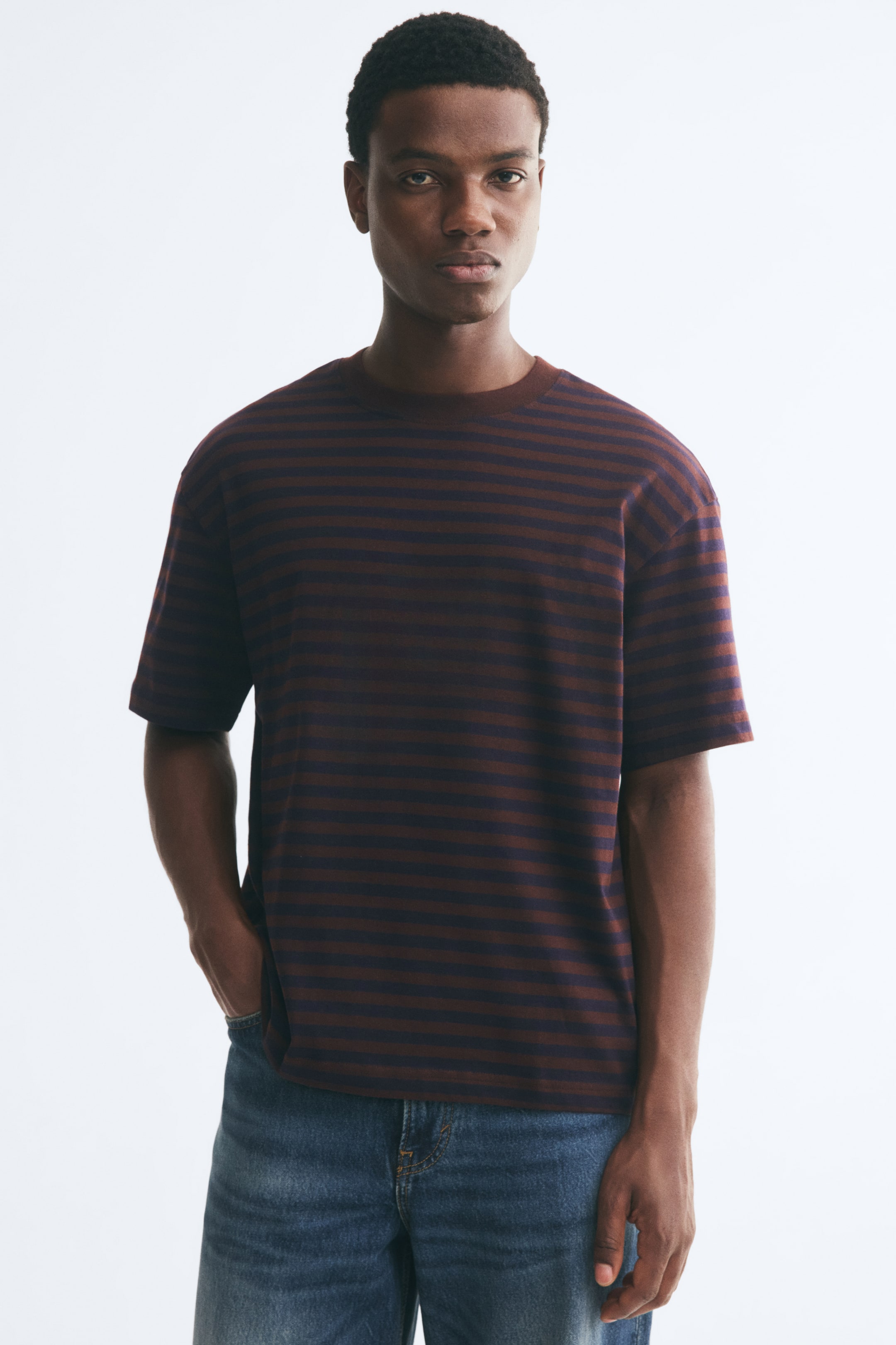 Agrandir l'image: Un t-shirt en coton rayé avec des bandes horizontales marron et bleu marine est porté par le mannequin. Le t-shirt présente une coupe décontractée avec des épaules tombantes, un col rond côtelé et un ourlet droit, associé à un jean bleu.