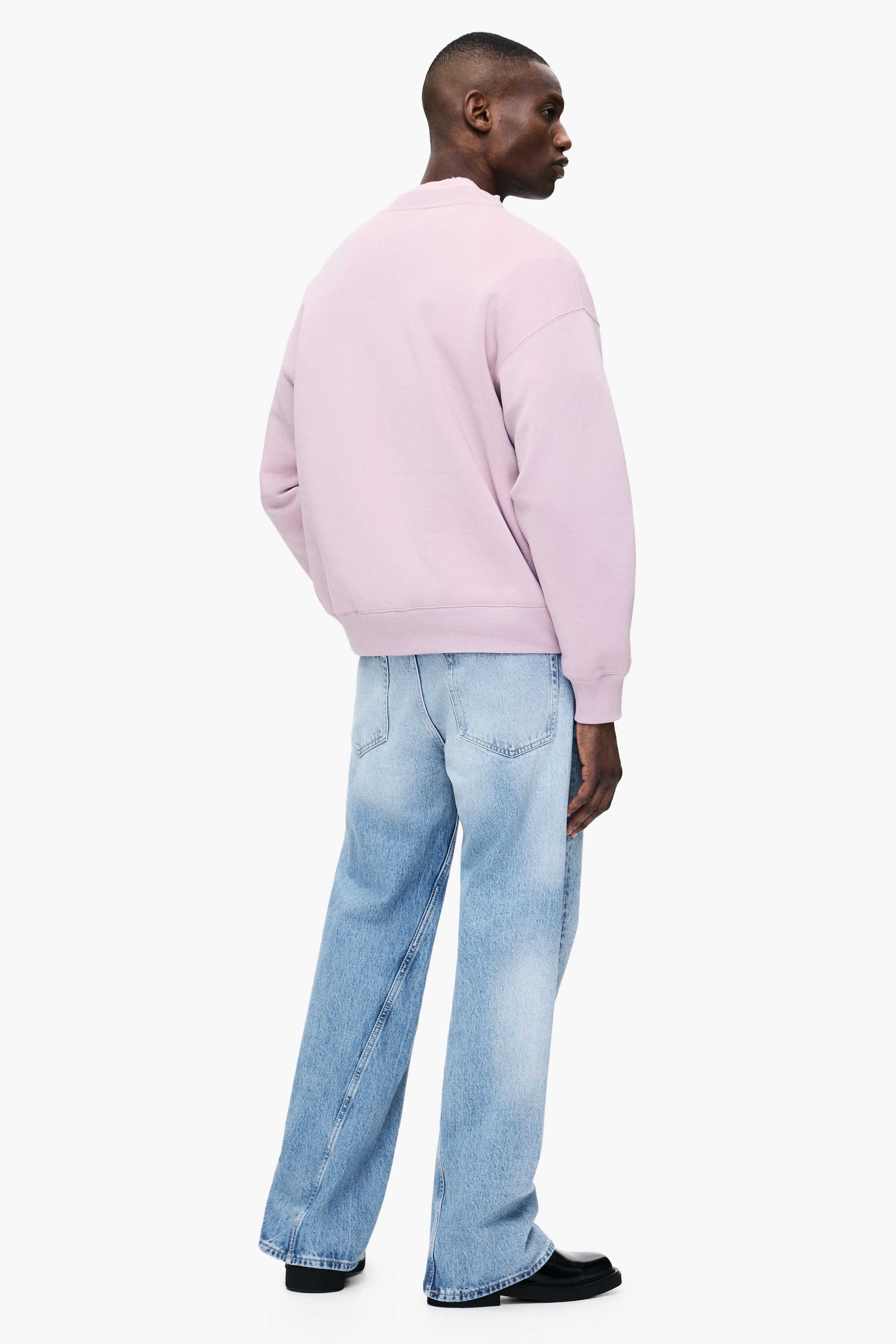 Zobrazit větší obrázek: Světlounce růžová mikina s kulatým výstřihem, která má volný střih s žebrovanými manžetami a spodním lemem, je nosena modelem. Model stojí s rukama v kapsách světlých džínů s širokými nohavicemi.
