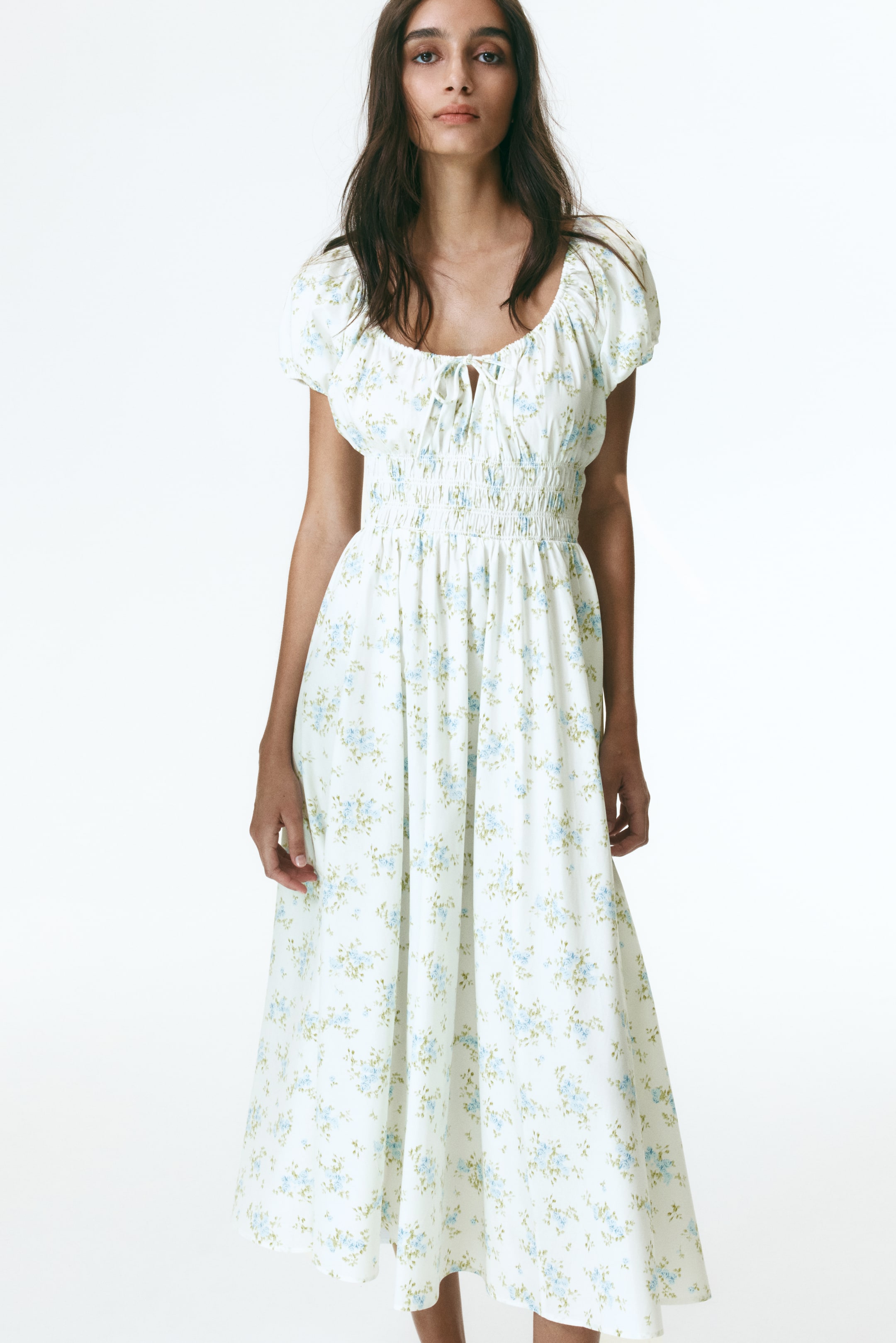 View larger image: The model is wearing a white midi dress adorned with a blue floral motif. It showcases a round, gathered neckline with a V-shaped opening and spaghetti ties at the front. The dress features short raglan puff sleeves, smocking at the waist, and a flared skirt.