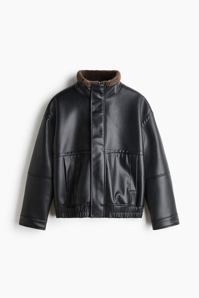 Eine schwarz beschichtete Stoffjacke ist von vorne zu sehen und zeigt einen hohen Stehkragen mit braunem Teddyfleece-Futter. Sie verfÃ¼gt Ã¼ber einen verdeckten FrontreiÃverschluss mit Windschutzleiste, seitliche Taschen und einen gesmokten elastischen Saum. - 1