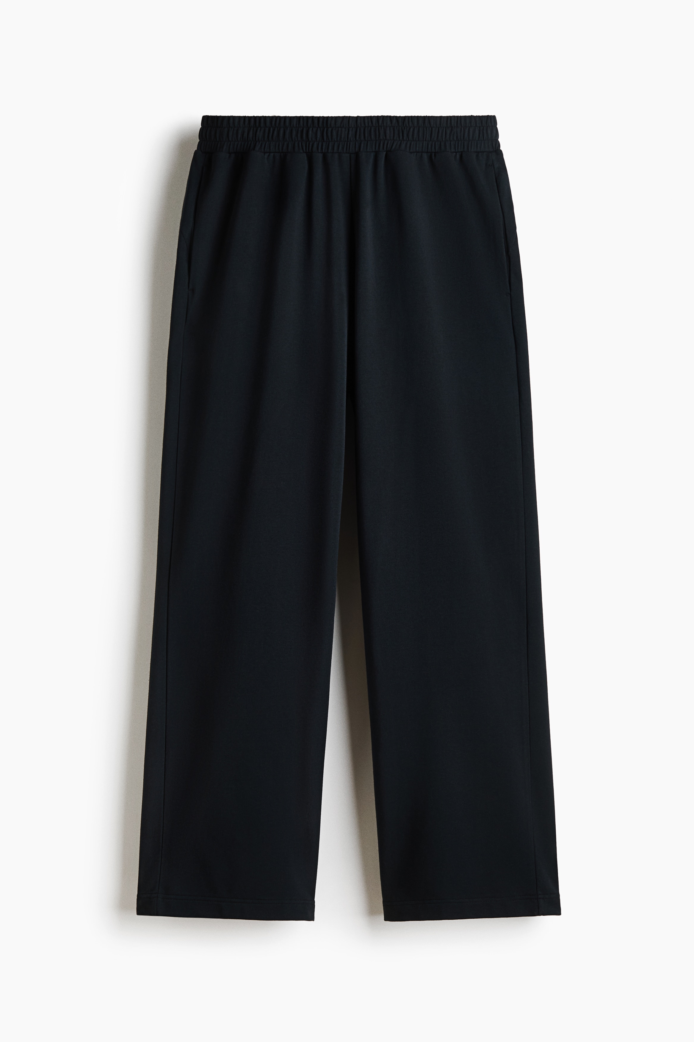 Black track pants with a loose-fit silhouette are worn by the model, who is standing with their hands in the side pockets. The pants are styled with a matching black short-sleeved T-shirt and black sneakers.