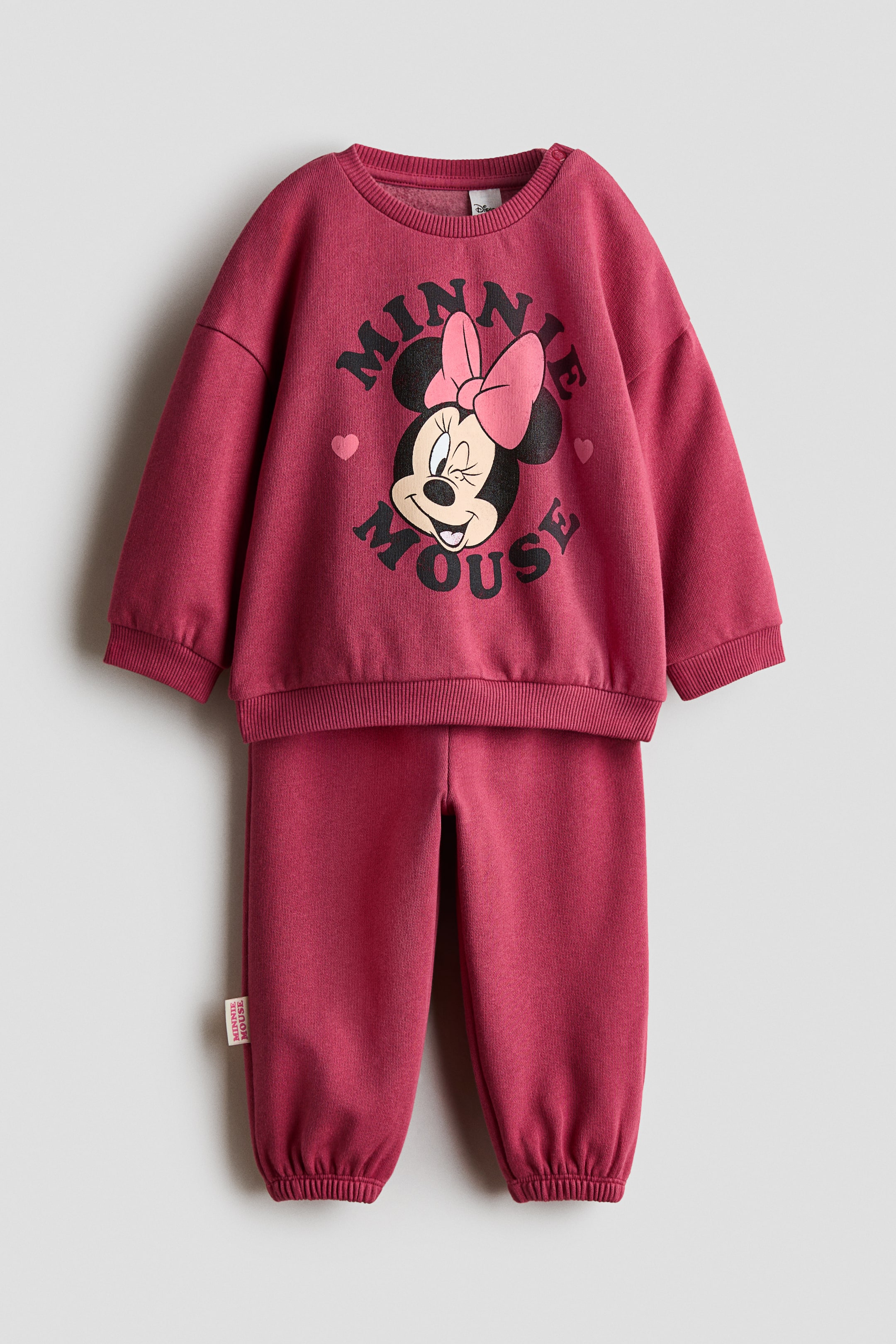 Ver imagen más grande: De frente, un chándal infantil de dos piezas en rosa baya presenta una sudadera de manga larga y un pantalón de chándal a juego. La sudadera muestra un cuello redondo acanalado, puños acanalados y un gráfico central de Minnie Mouse guiñando un ojo con el texto “MINNIE MOUSE” arqueado a su alrededor. El pantalón de chándal a juego presenta un corte holgado con bajos elásticos en los tobillos.