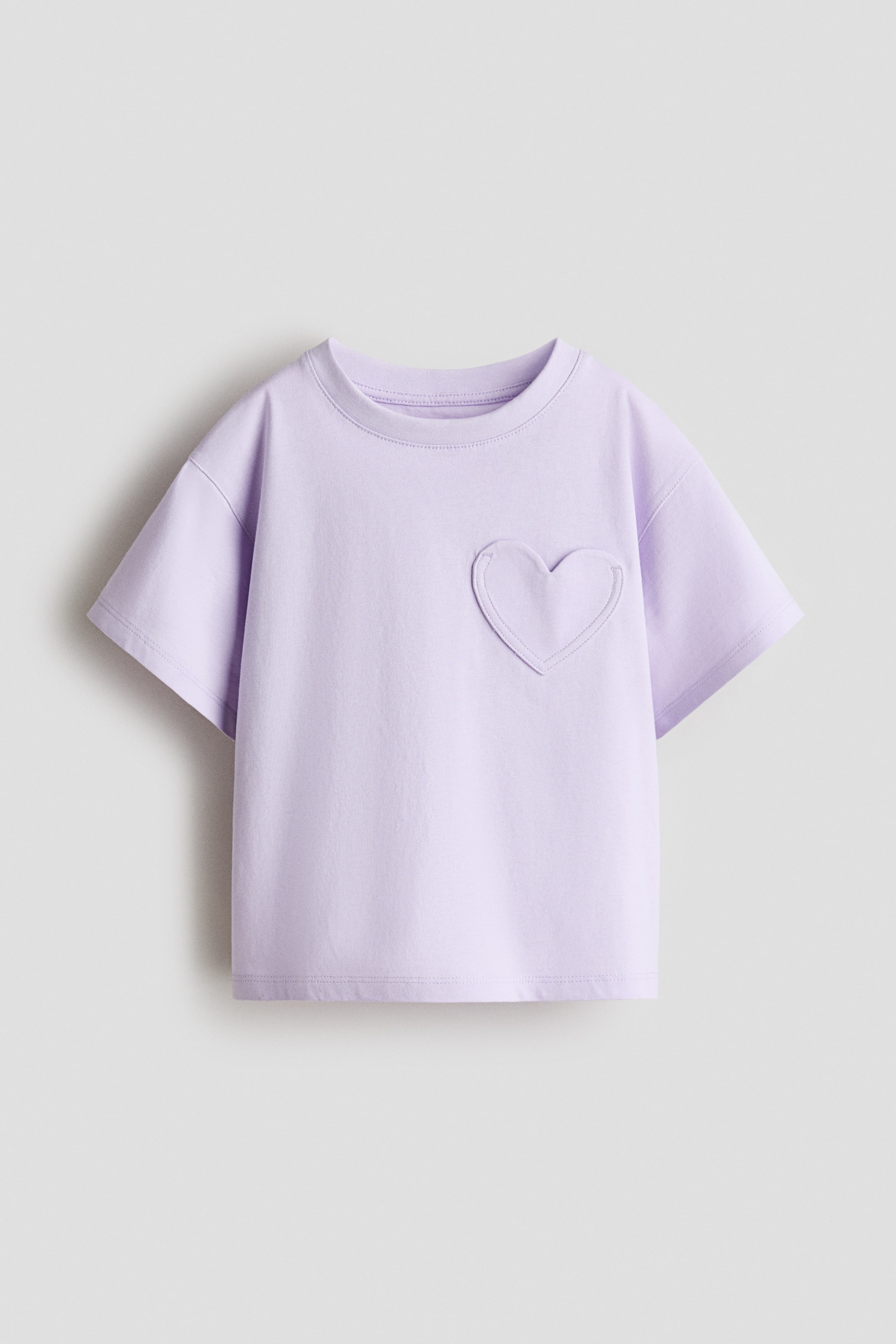 拡大画像を表示: ライトパープルの半袖Tシャツが前を向いており、リブ付きのクルーネックが特徴です。左胸にはハート型のパッチが縫い付けられています。