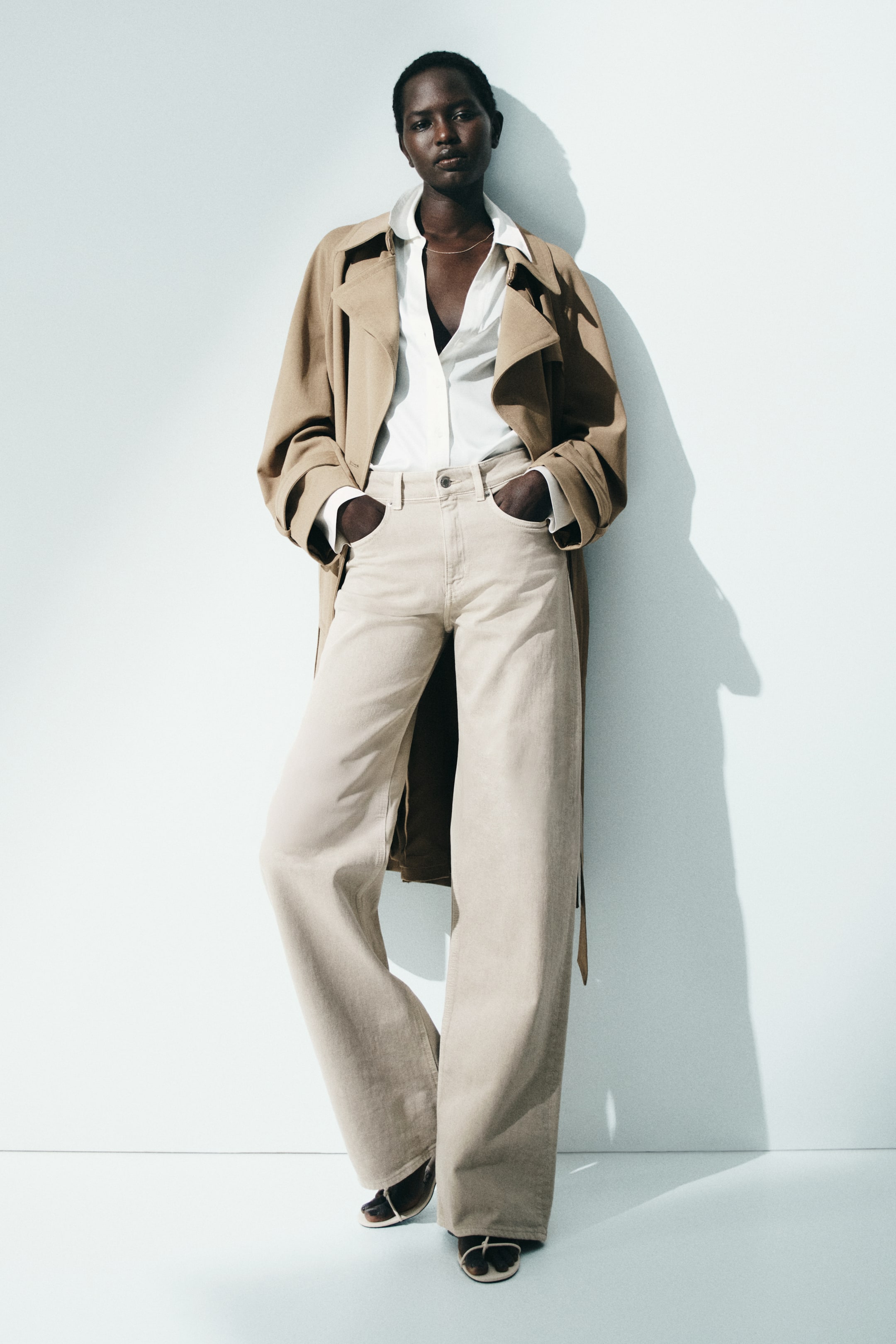 View larger image: A layered ensemble showcases a light brown trench coat with wide lapels and pushed-up sleeves, worn open over a white unbuttoned shirt and light beige wide-leg trousers. The trousers feature visible front pockets and belt loops, while dark open-toed sandals complete the look at the feet.