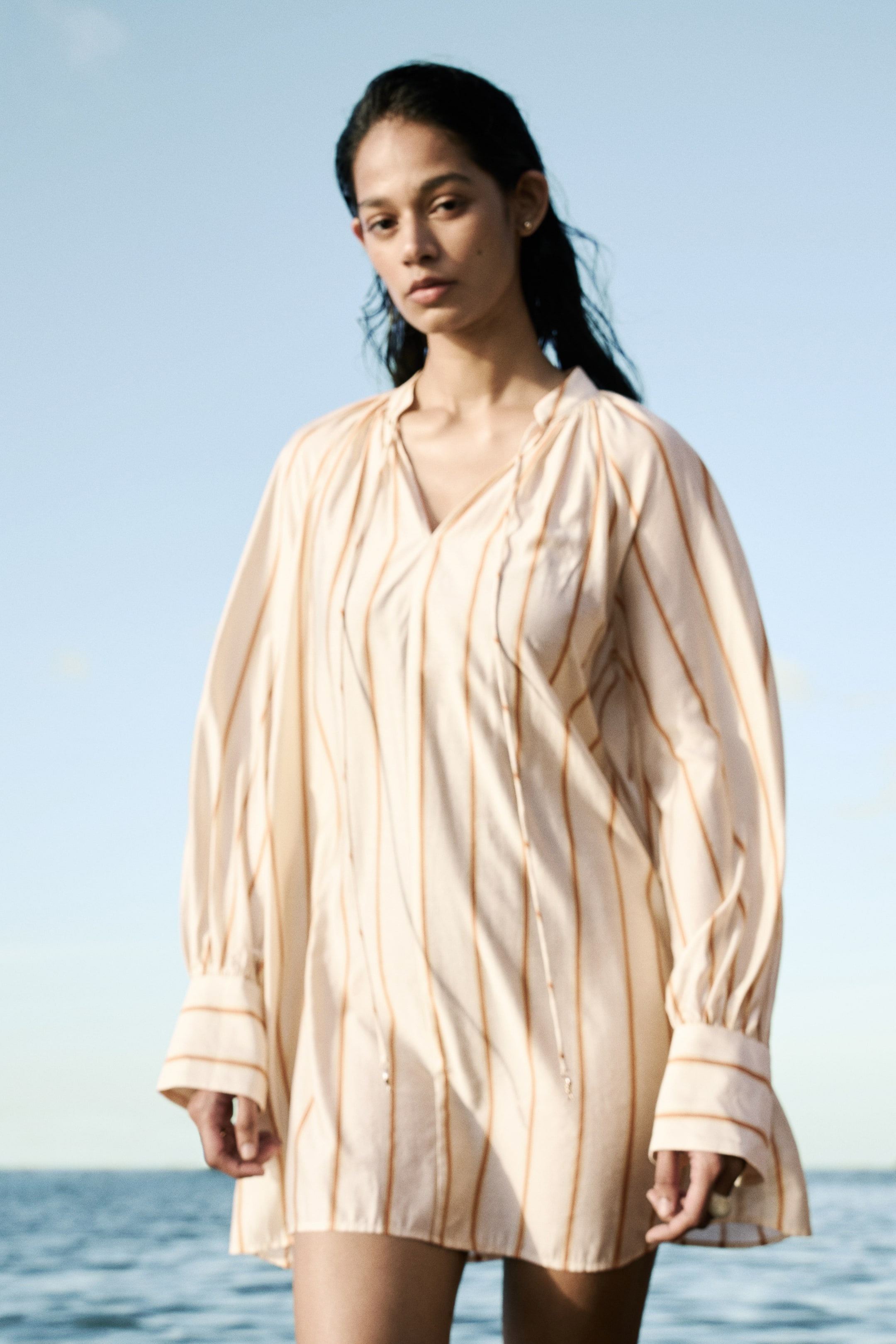 View larger image: An off-white dress showcasing thin vertical orange stripes. Designed with a V-neckline and tie closures, its long, voluminous sleeves gather into wide cuffs.