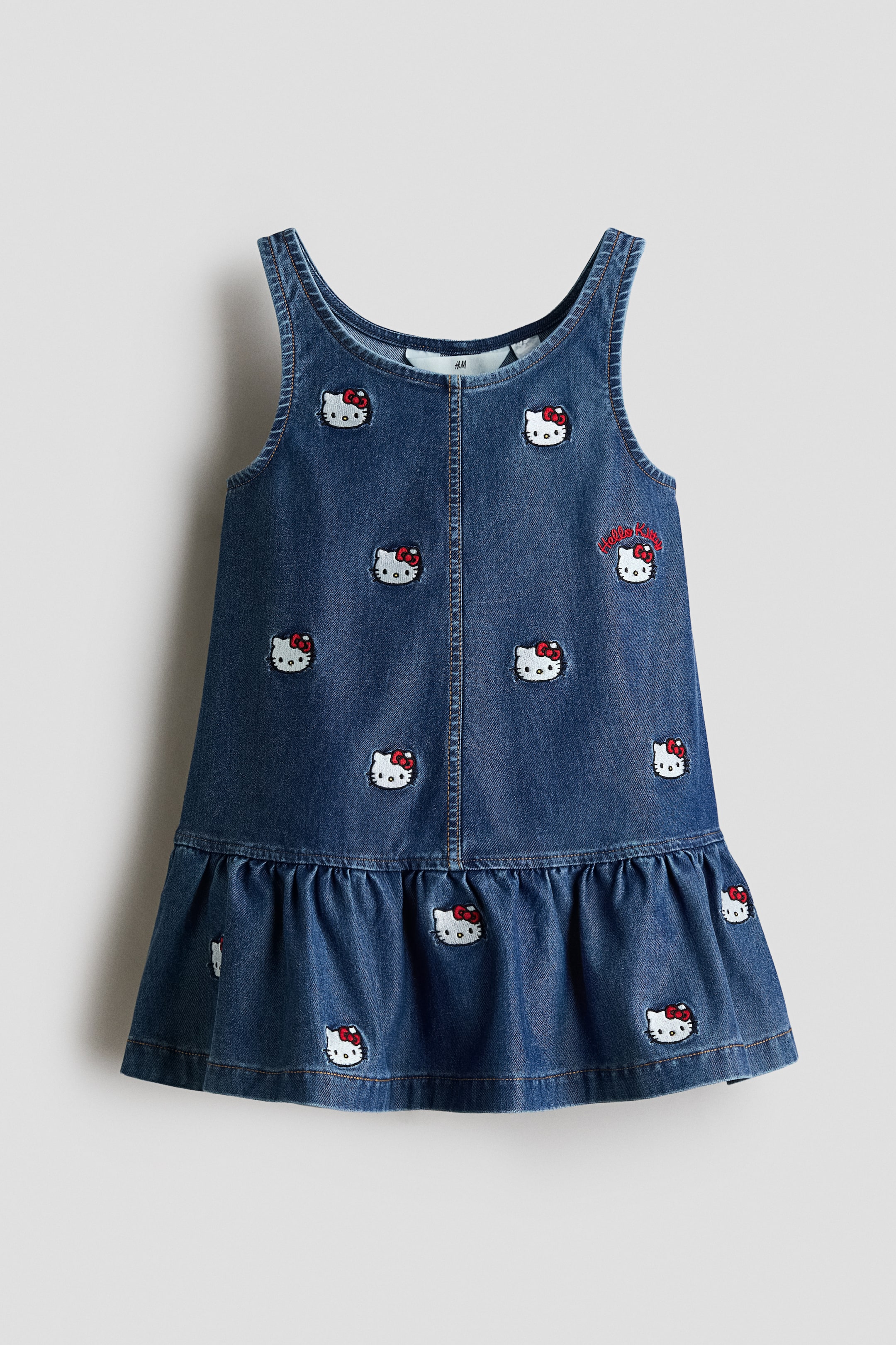 View larger image: A blue denim dress is showcasing its front view, highlighting a sleeveless design with broad straps and a scoop neckline. It is embellished with multiple embroidered Hello Kitty faces adorned with red bows and a 'Hello Kitty' text embroidery on the upper right chest, complemented by a gathered ruffle forming the hem.