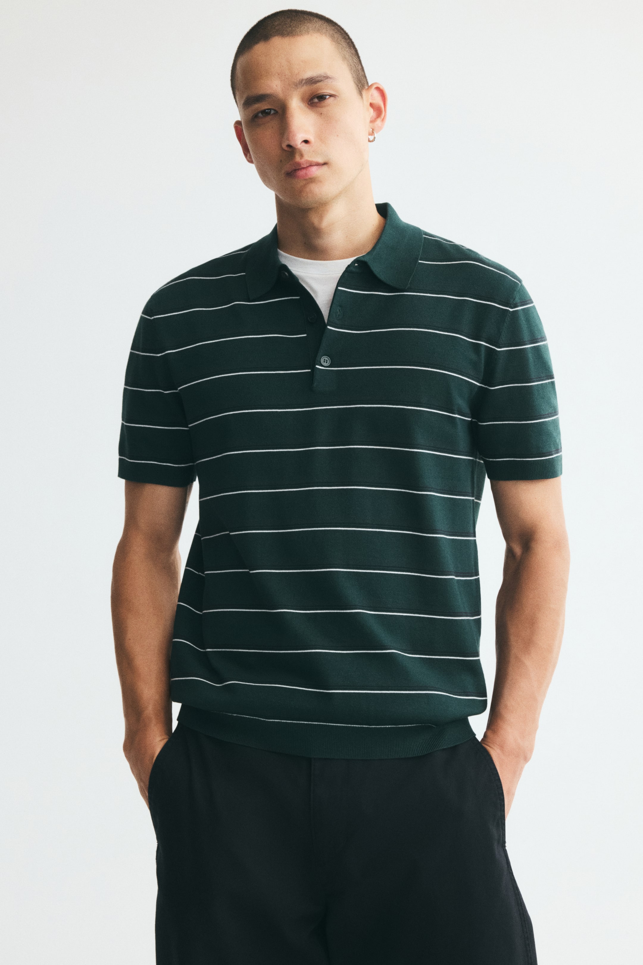 View larger image: A green knit polo shirt with thin horizontal white stripes is worn by the model over a white crew-neck T-shirt. The shirt features a classic collar, a partially open button placket, and short sleeves, and is styled with black trousers.