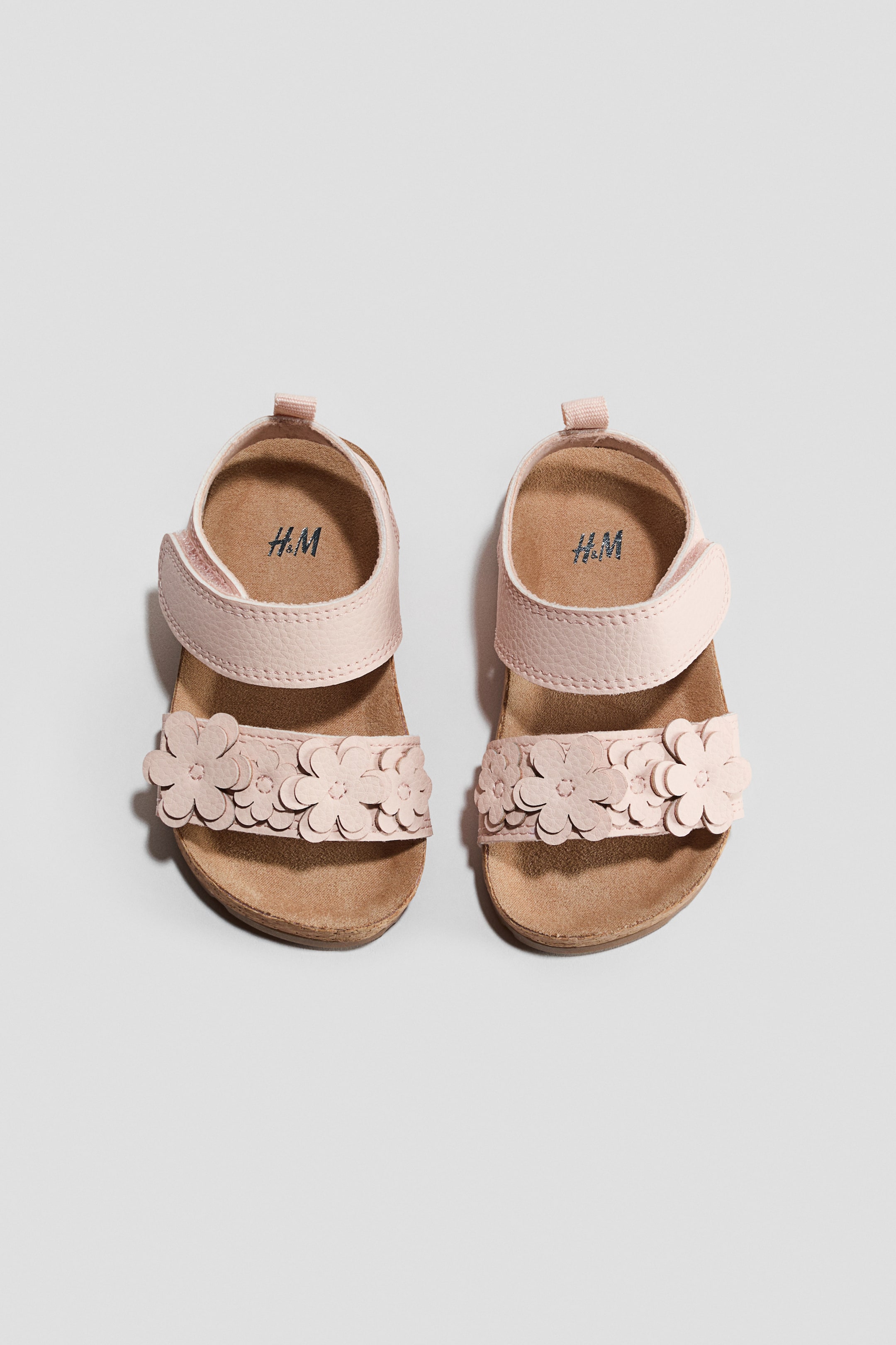 View larger image: Introducing a pair of light pink sandals showcased from the front, featuring a foot strap embellished with floral appliques and an ankle strap, both secured with a hook and loop fastening. These sandals boast moulded insoles and fluted soles, perfect for adding a touch of elegance to any outfit.