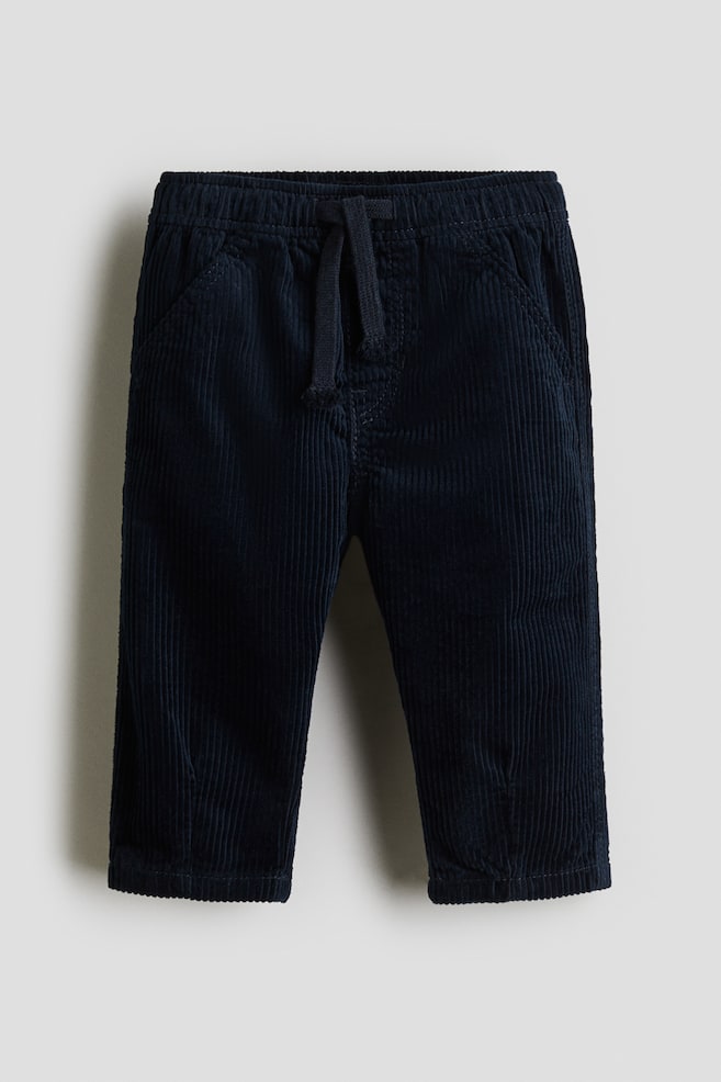 A front view of navy corduroy pants shows an elasticated waistband with a tied drawstring and two front slash pockets. The pants feature a distinct vertical corduroy texture. - 1