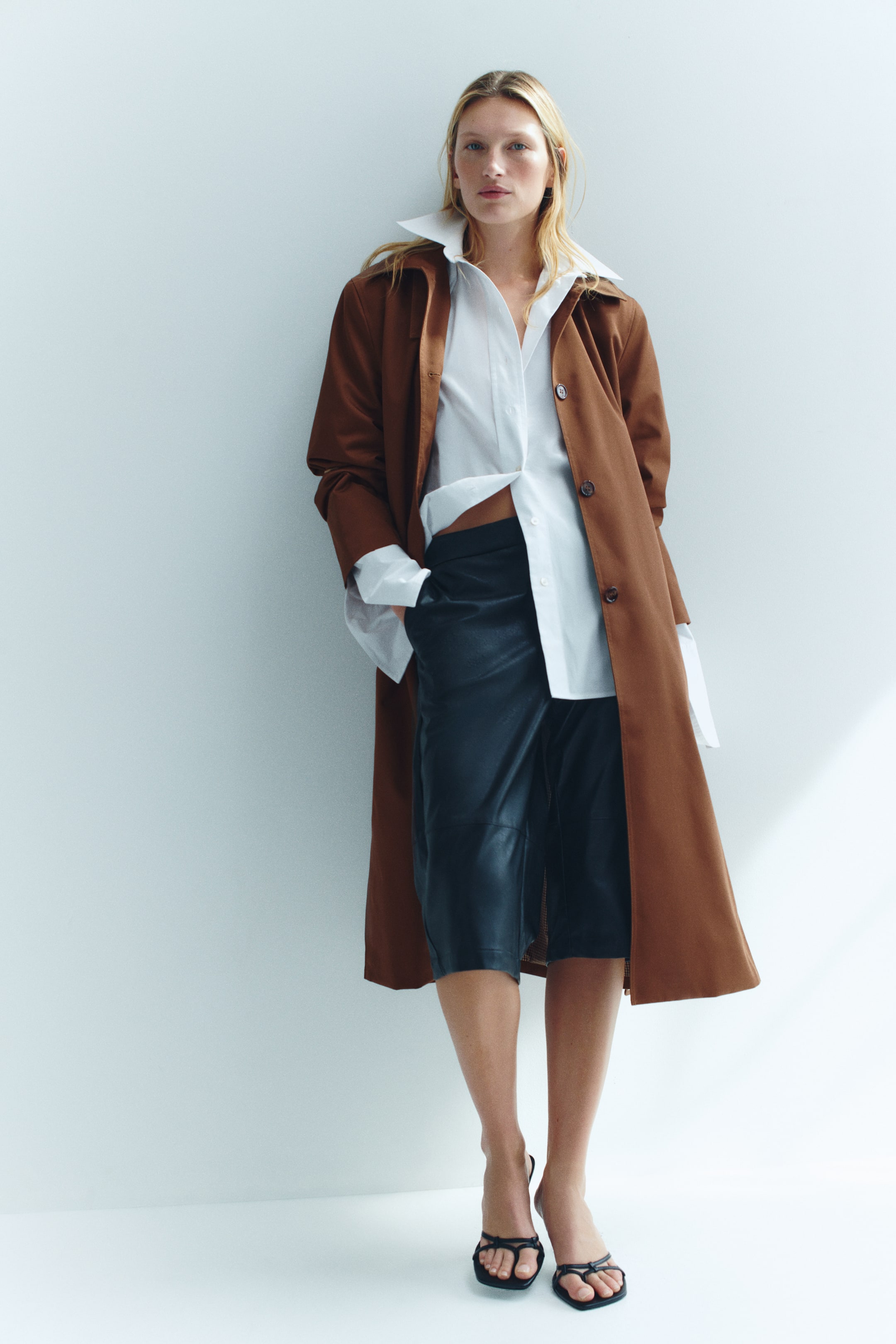 Visualizza immagine più grande: Un car coat oversize in twill marrone, lungo fino a metà polpaccio, indossato aperto dalla modella, con colletto classico e chiusura frontale con bottoni. Il cappotto è abbinato a una camicia bianca con bottoni e a pantaloncini scuri al ginocchio.