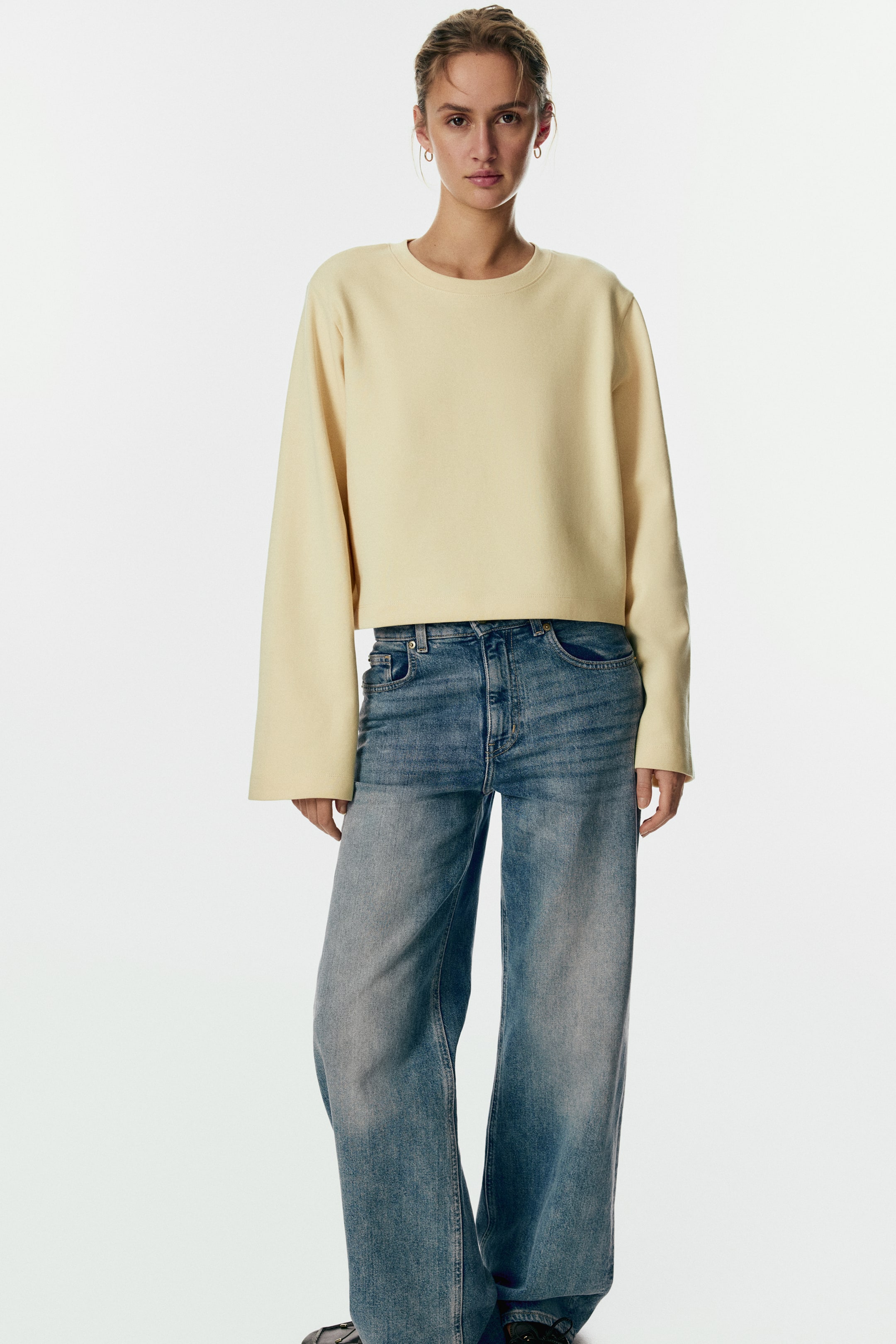 Größeres Bild anzeigen: Ein blasses gelbes Sweatshirt mit Rundhalsausschnitt wird getragen, das einen entspannten, leicht verkürzten Schnitt, lange weite Ärmel ohne Bündchen und einen geraden Saum aufweist.