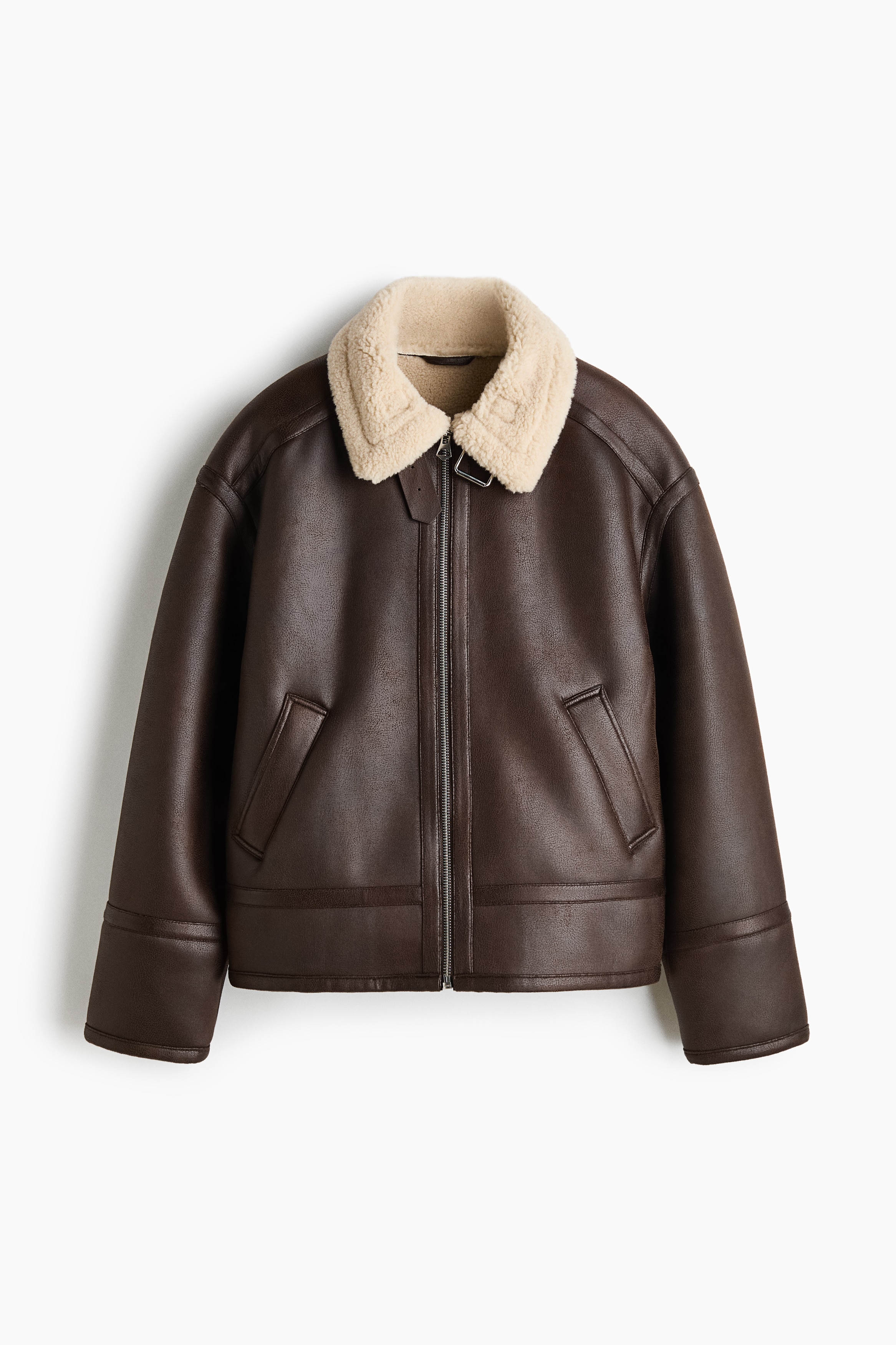 A dark brown jacket in coated fabric with a soft cream-coloured teddy collar and lining is being worn open by the model. The jacket features a front metal zipper, an adjustable throat latch with a metal buckle, and welt pockets, layered over a blue checkered shirt and light-coloured trousers.